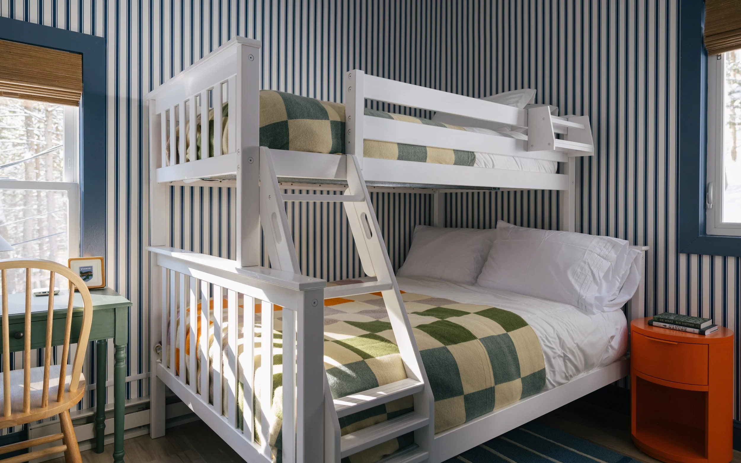 Spacious bunk room at Pine & Plaid Vermont cabin with bunk bed over full bed, striped wallpaper, cozy workspace, and winter views near The Quechee Club.