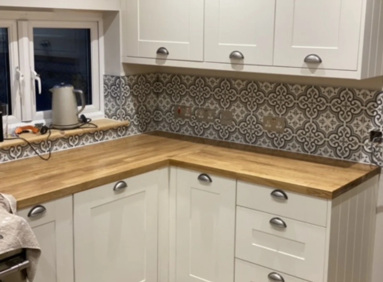 A kitchen corner with white cabinets, a patterned backsplash, and a wooden countertop. There is a window with a kettle and an orange-handled tool on the windowsill, and electrical outlets on the backsplash.