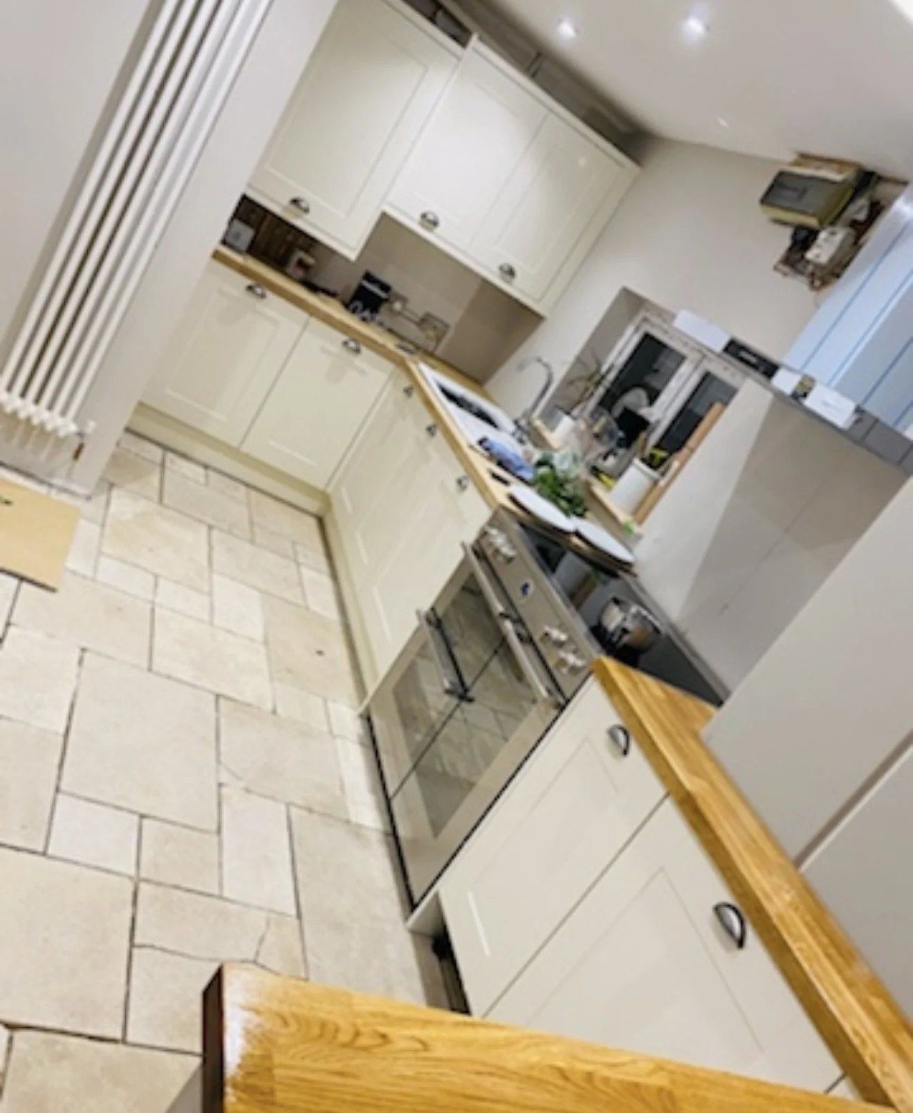 A kitchen with cream-colored cabinets, a wooden countertop, a stove, and a window above the sink. There are various items on the counter including a plant and dishes.
