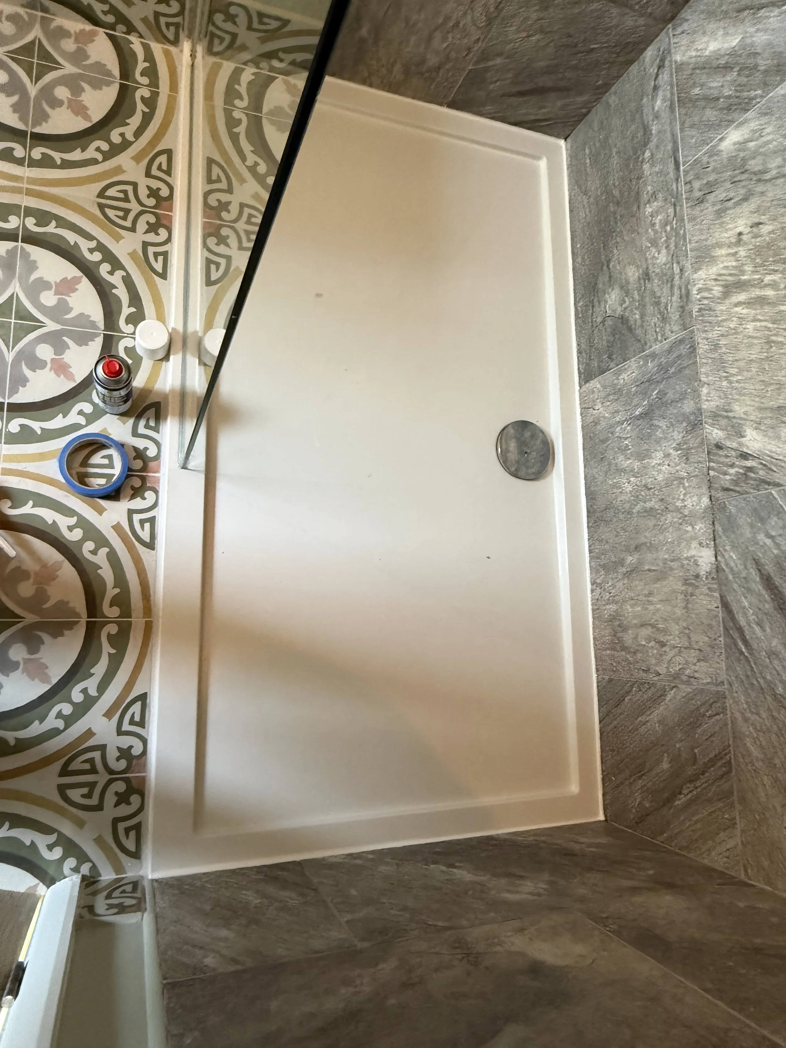 Empty shower stall with a white base and a small round drain on the right side, surrounded by decorative patterned tiles and gray tiled walls.
