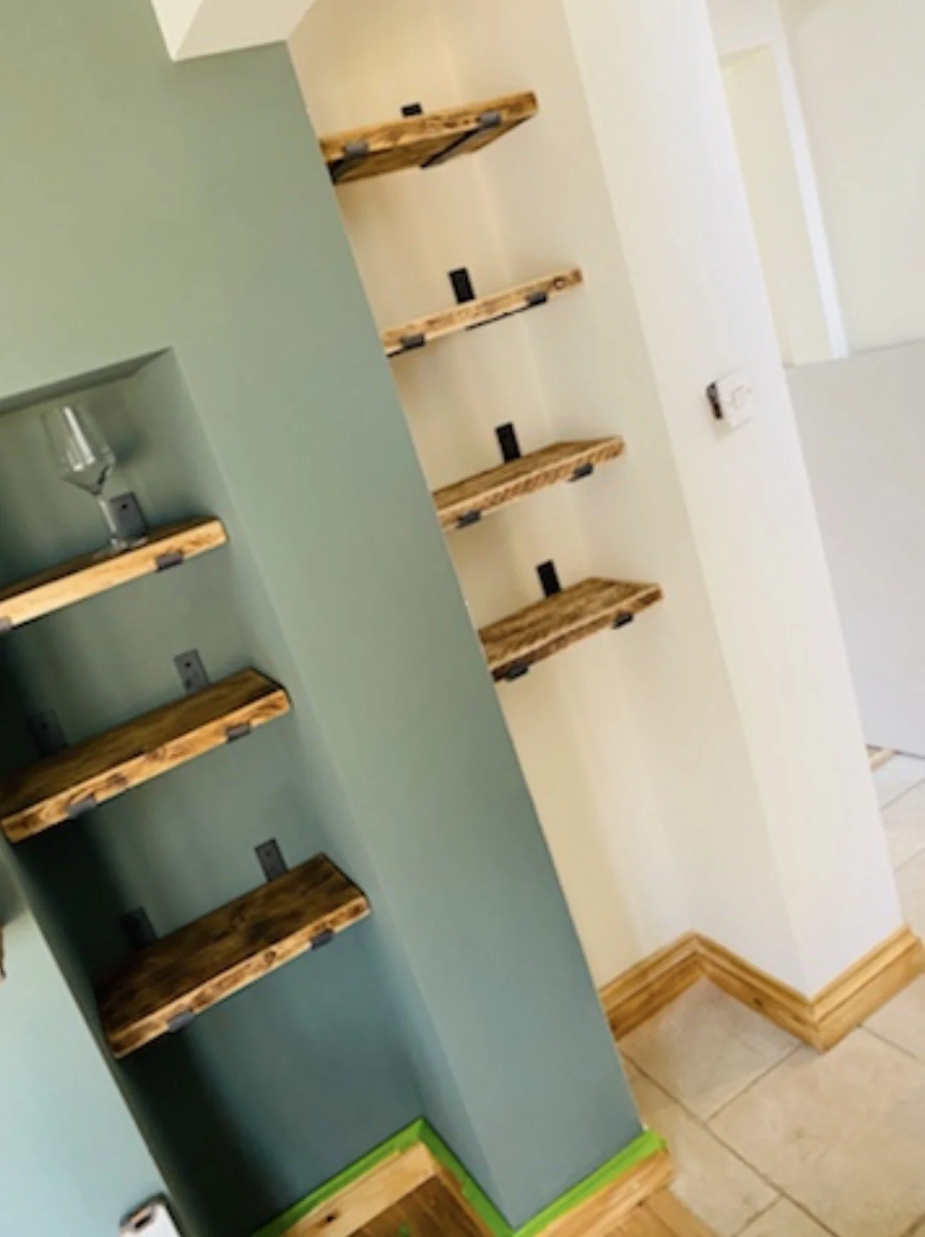 Close-up of a wall with wooden floating shelves against a green-painted wall, with an adjacent cream-colored wall and floor tiles.
