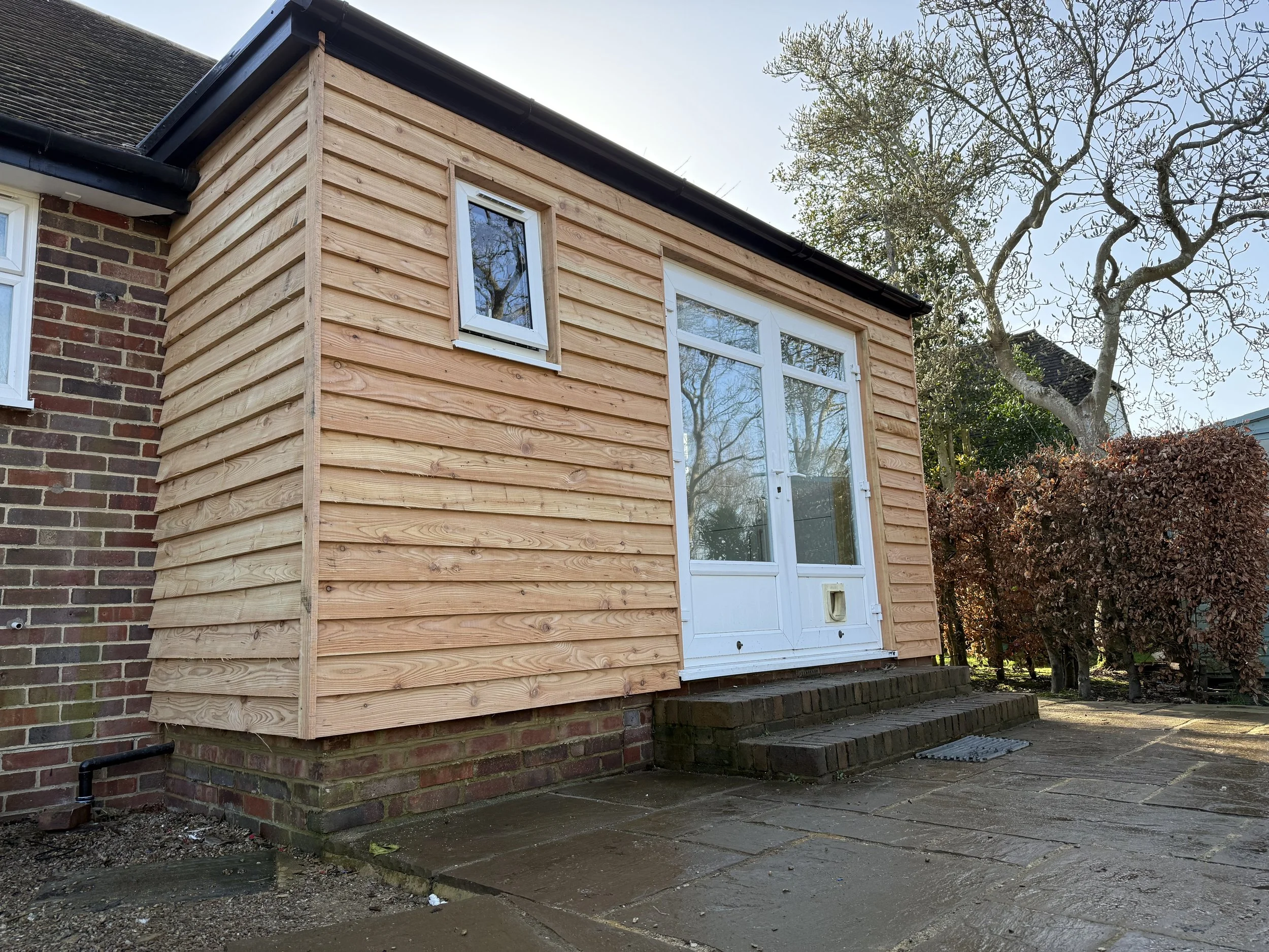A small extension with wooden cladding added to the back of a brick house, featuring a glass door and a small window, with steps leading up to the door and a pathway in front.