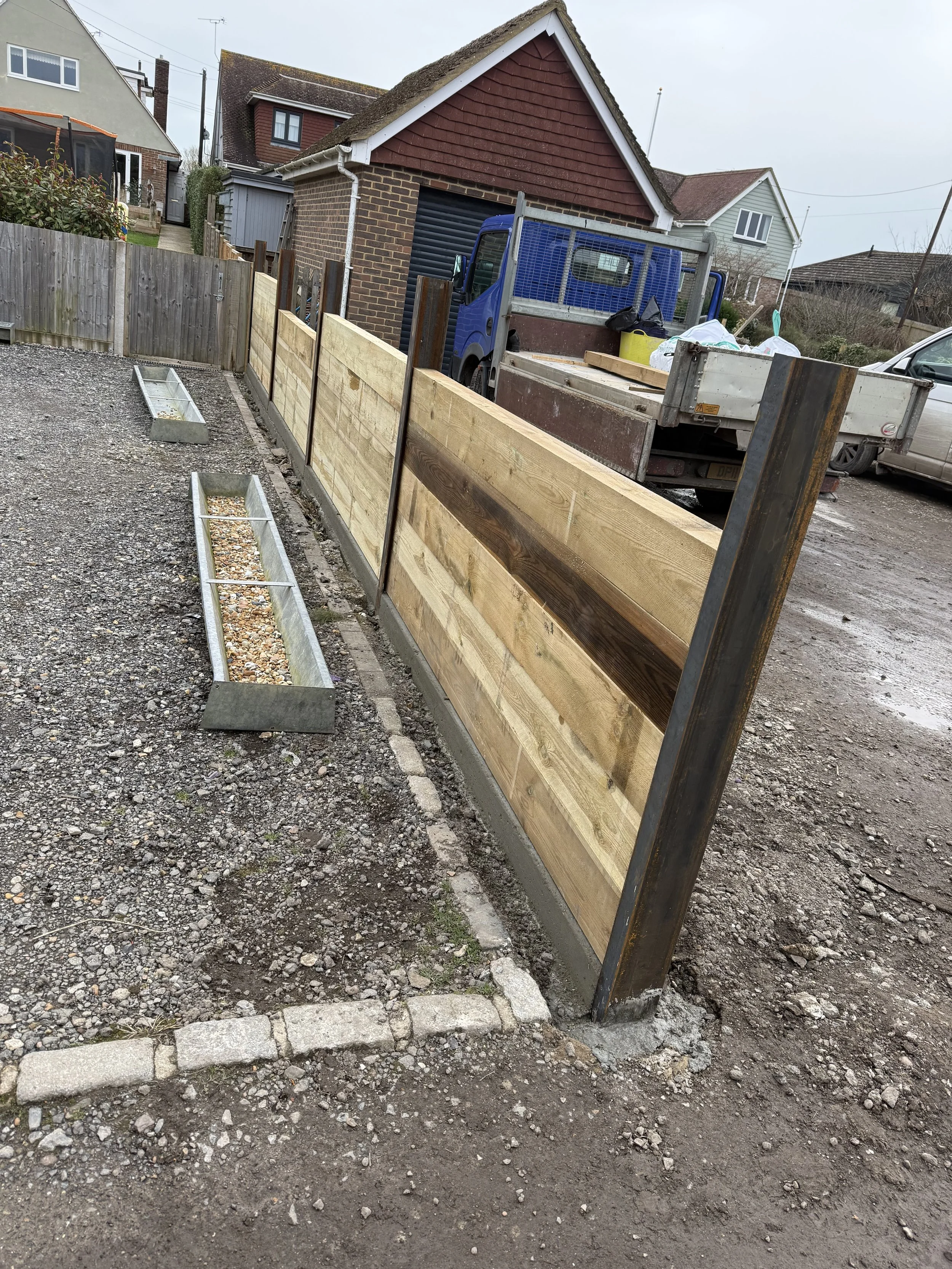 A newly built wooden fence with metal posts along a gravel driveway in a residential area, with a row of rectangular flower beds filled with gravel or small rocks in front of it, and houses and a truck in the background.