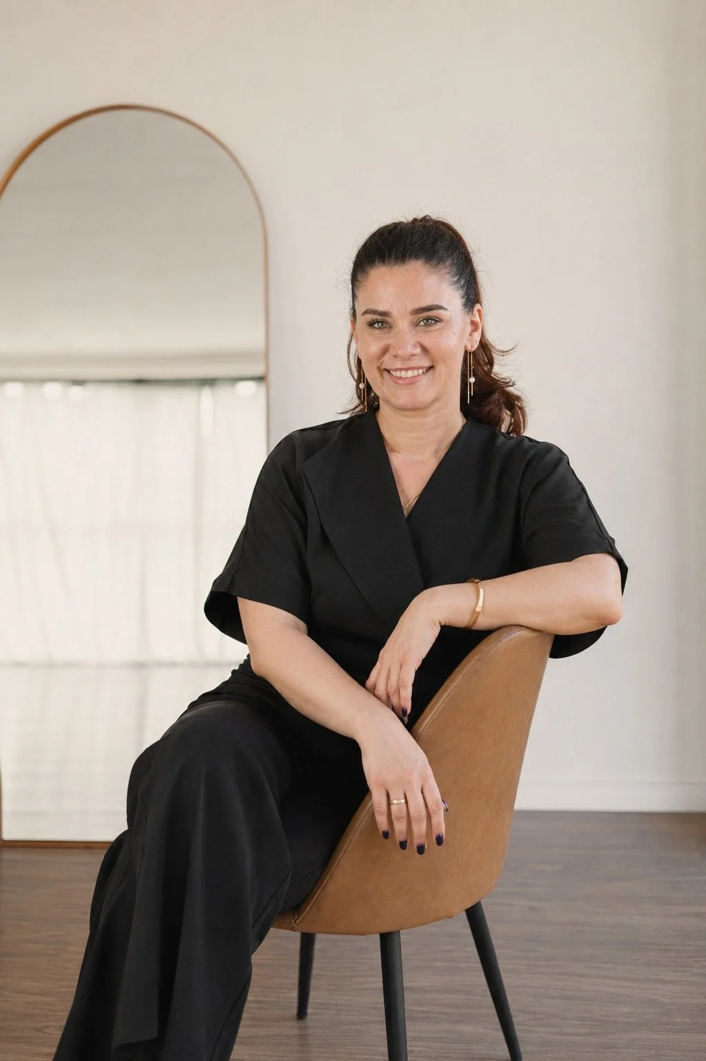 A woman with dark hair tied back, wearing a black outfit, sitting on a tan chair in a room with beige walls, smiling at the camera with a mirror behind her.