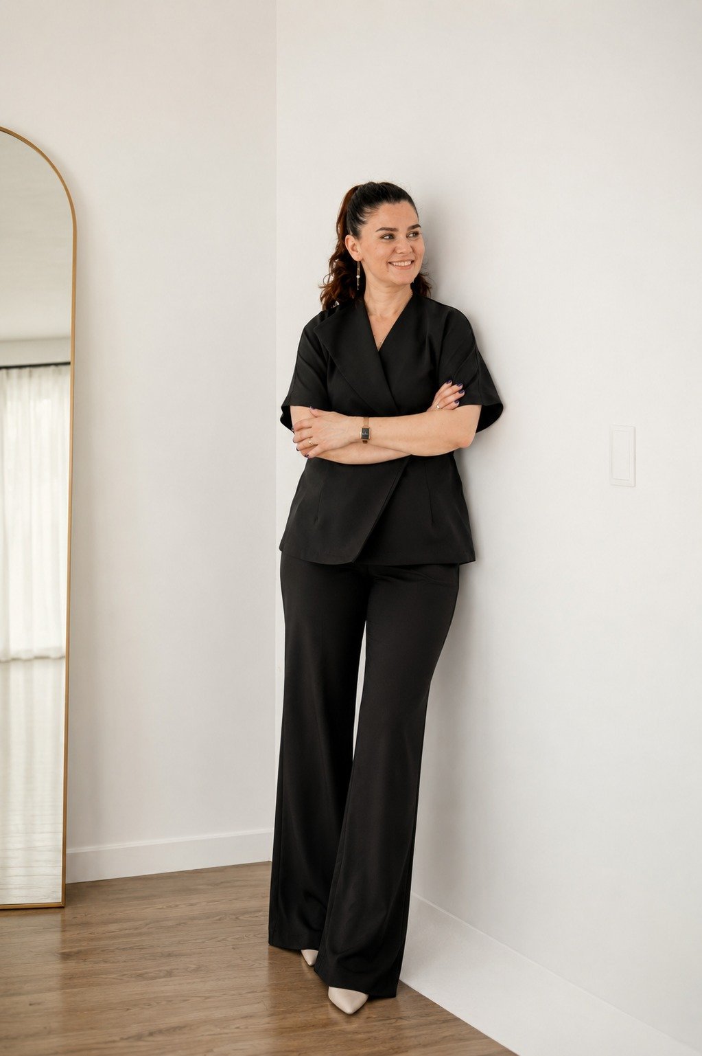 A woman in a black suit with wide-leg pants and a blazer, standing with her arms crossed and smiling, in a room with a white wall and a mirror.