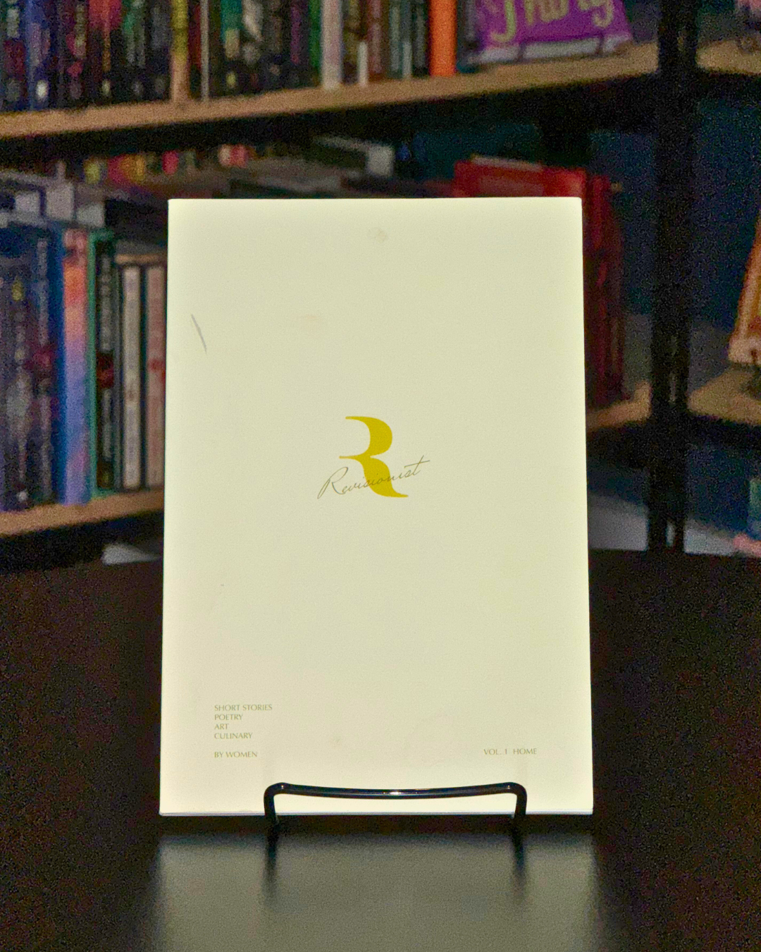 A white magazine titled 'Remindut' with a golden crescent moon symbol and the words 'SHORT STORIES, POETRY, ART, CULINARY, BY WOMEN, VOL. 1 HOME' on a black stand, set against bookshelves filled with colorful books.
