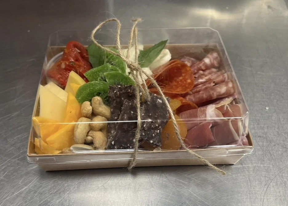 Charcuterie board with various cheeses, cured meats, dried fruits, nuts, and fresh vegetables in a clear plastic box tied with twine.