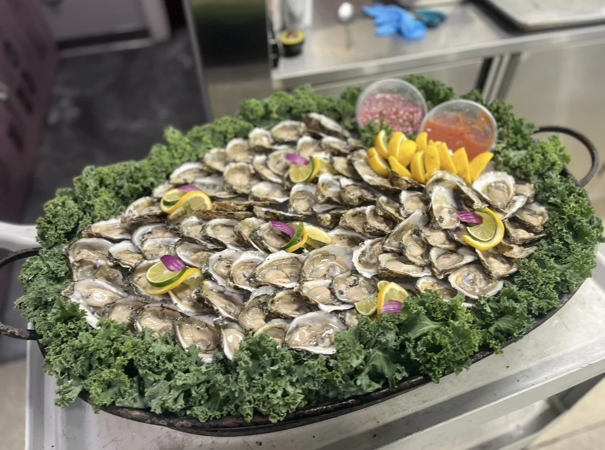 A tray of raw oysters served on ice, garnished with lemon slices, purple onion slices, and parsley, with two small containers of cocktail sauce and mignonette sauce in the background.