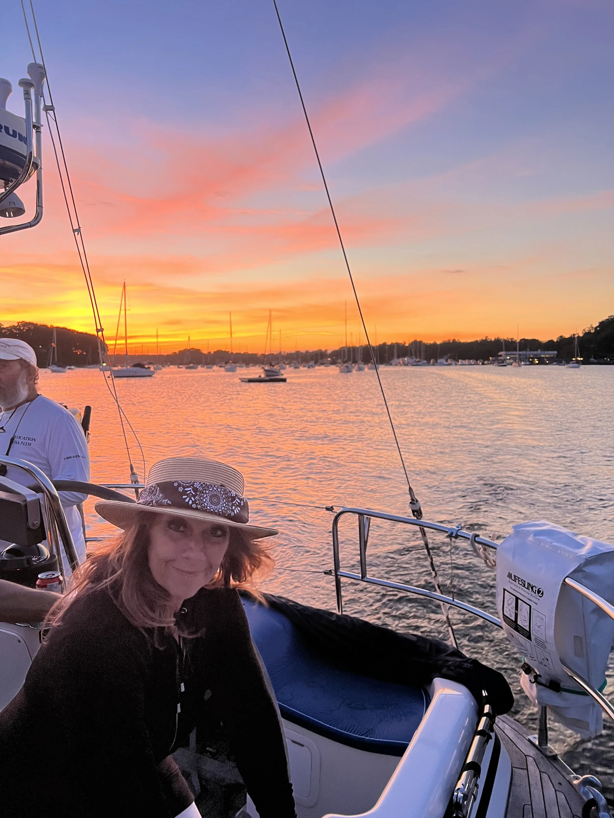 A woman with light brown hair and a wide-brimmed hat smiling on a boat during sunset with orange, pink, and purple sky, and other boats floating on the water.