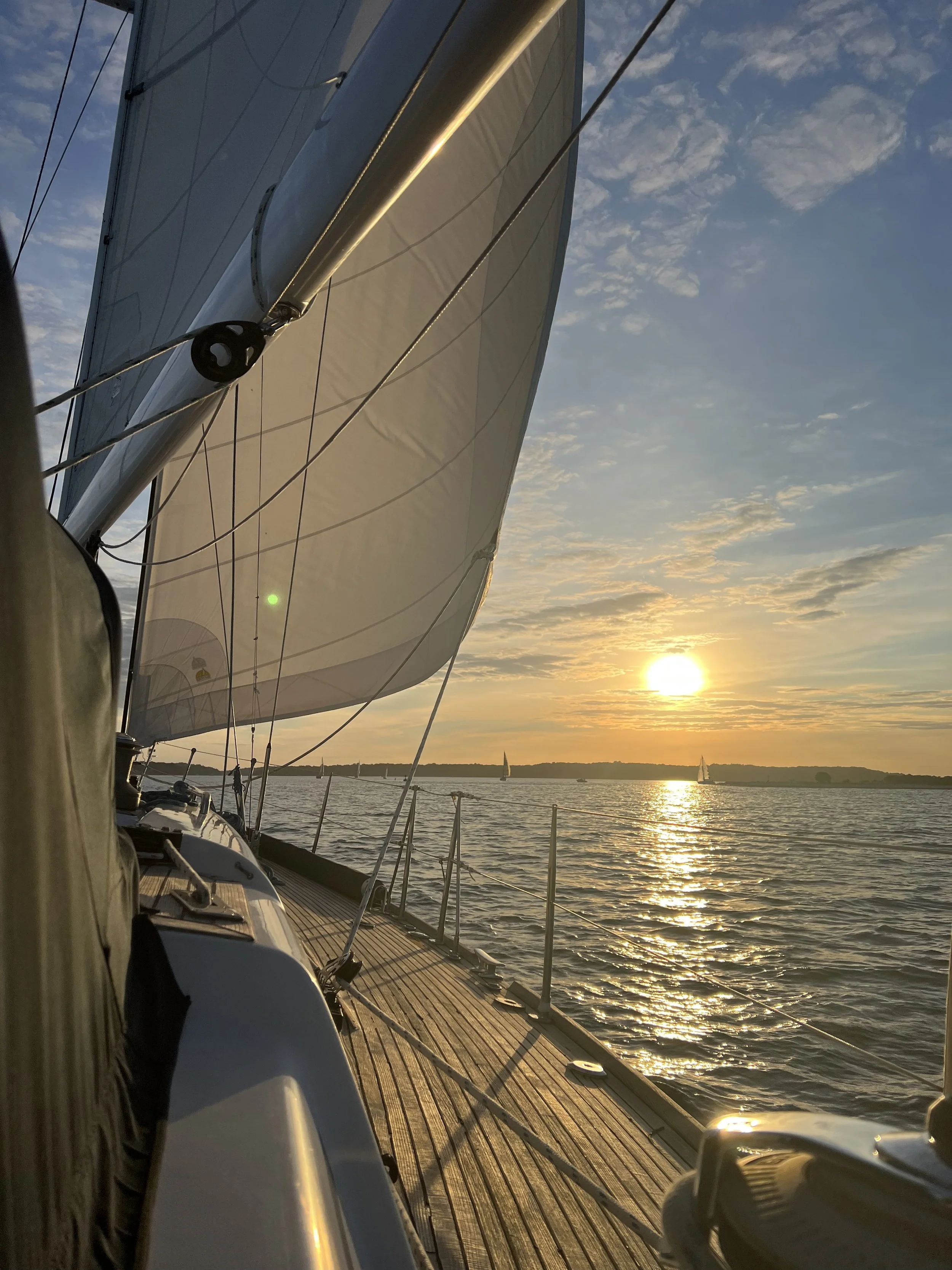 A sailboat deck during sunset with the sail up and the sun reflecting on calm water, with other sailboats visible in the distance.