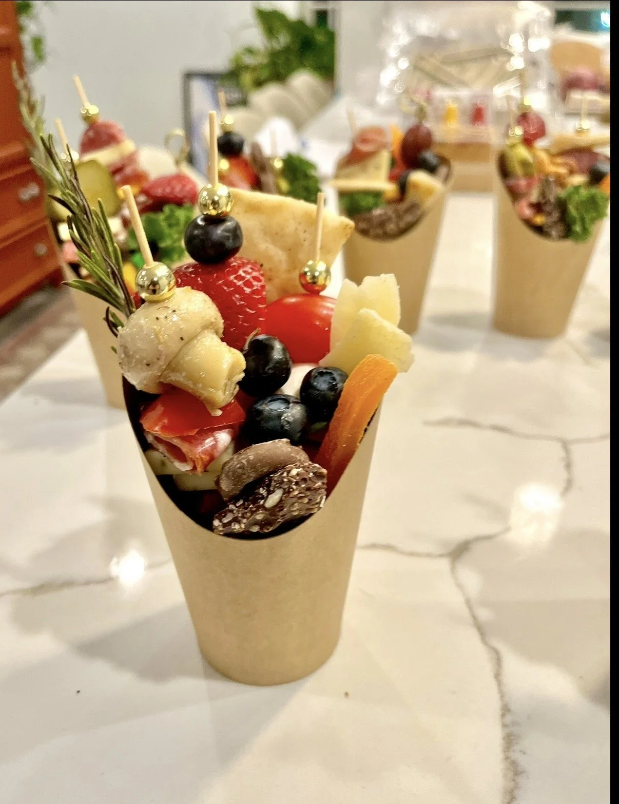 A charcuterie cone filled with assorted cheeses, fruits, nuts, and herbs, garnished with small decorative gold balls and a sprig of rosemary.