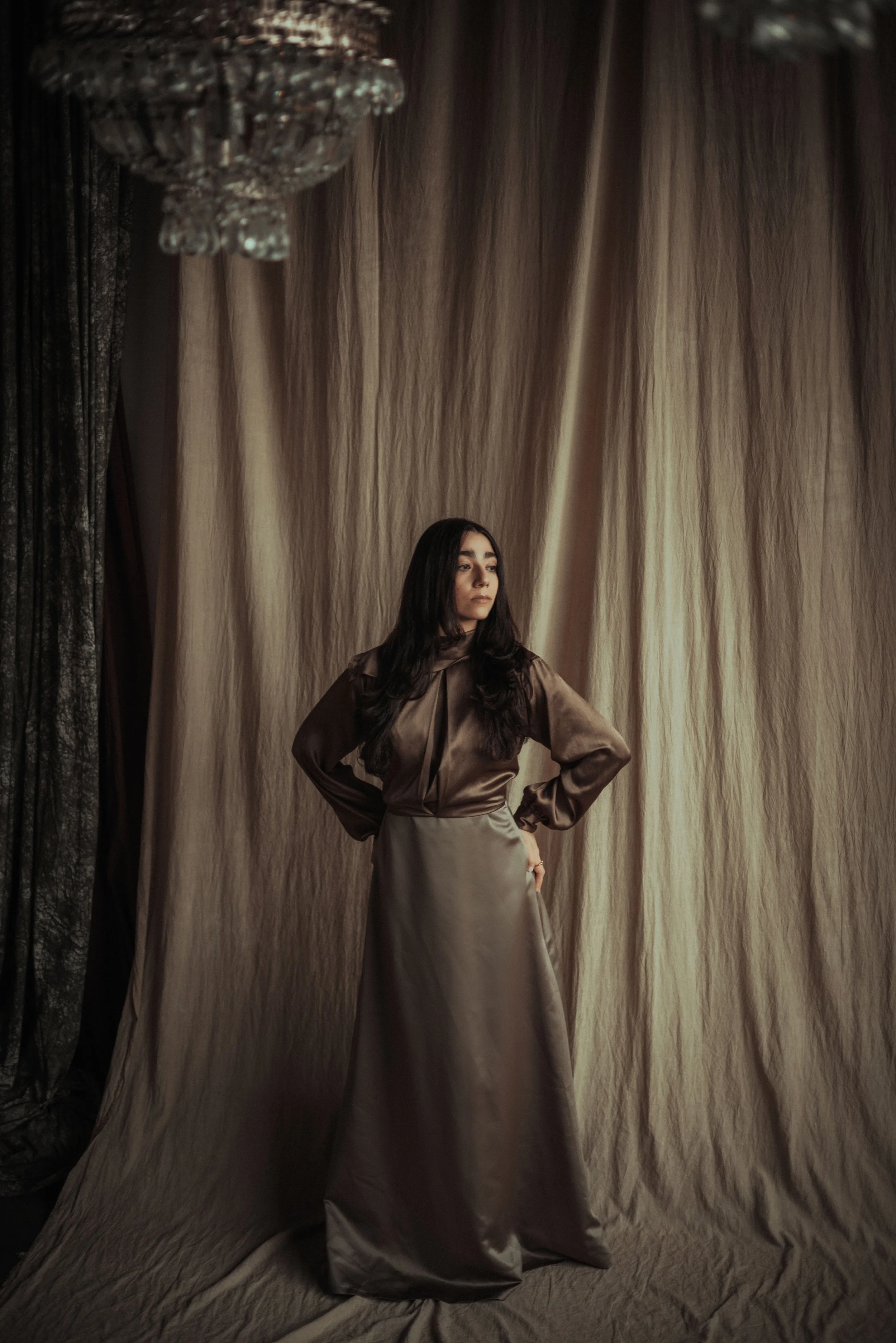 A woman with long dark hair wearing a shiny brown blouse and a beige satin long skirt stands with her hands on her hips in front of beige curtains and a crystal chandelier.