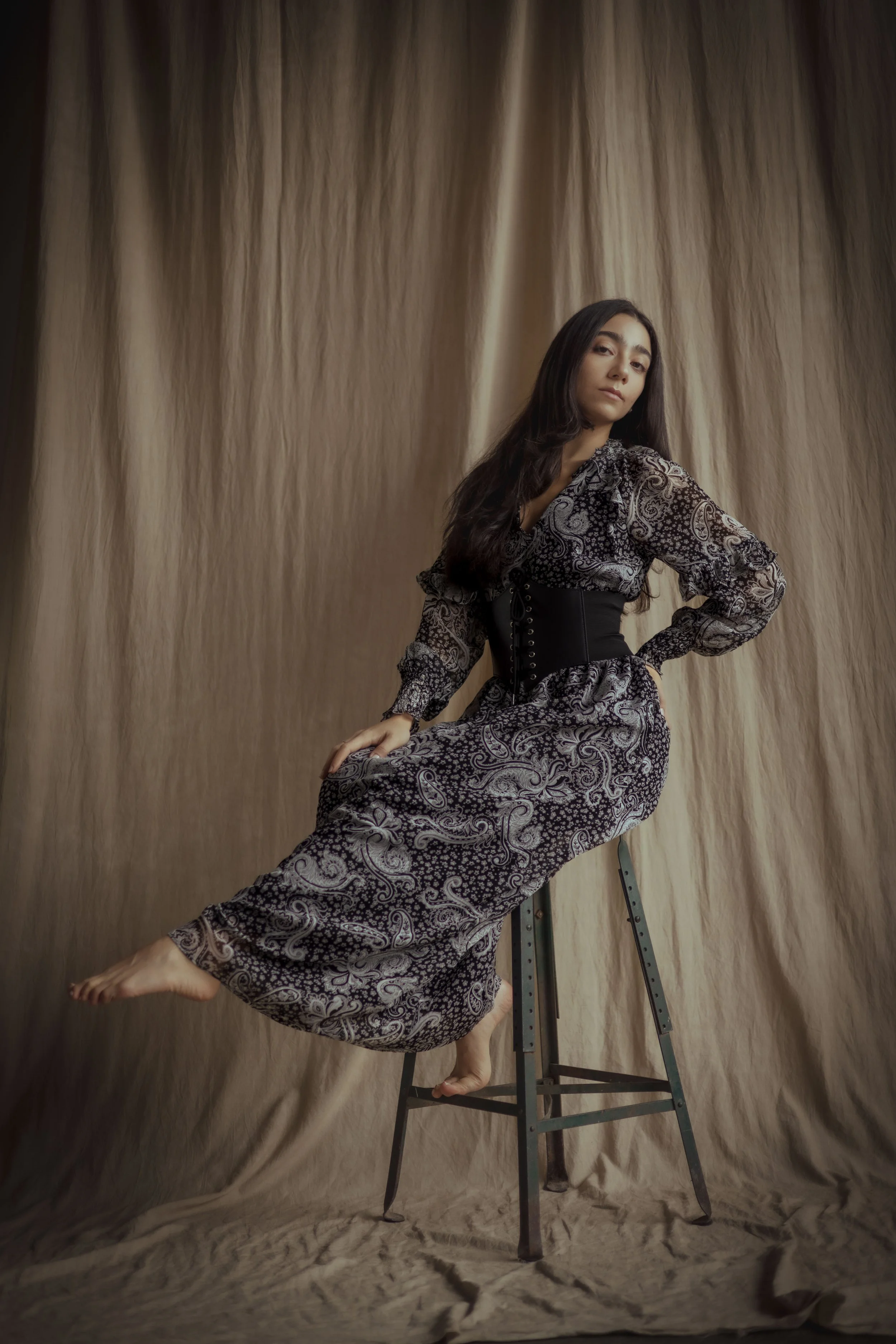 A woman with long dark hair seated on a black step stool in front of a beige fabric backdrop, wearing a patterned dress with a black waist corset, looking at the camera.