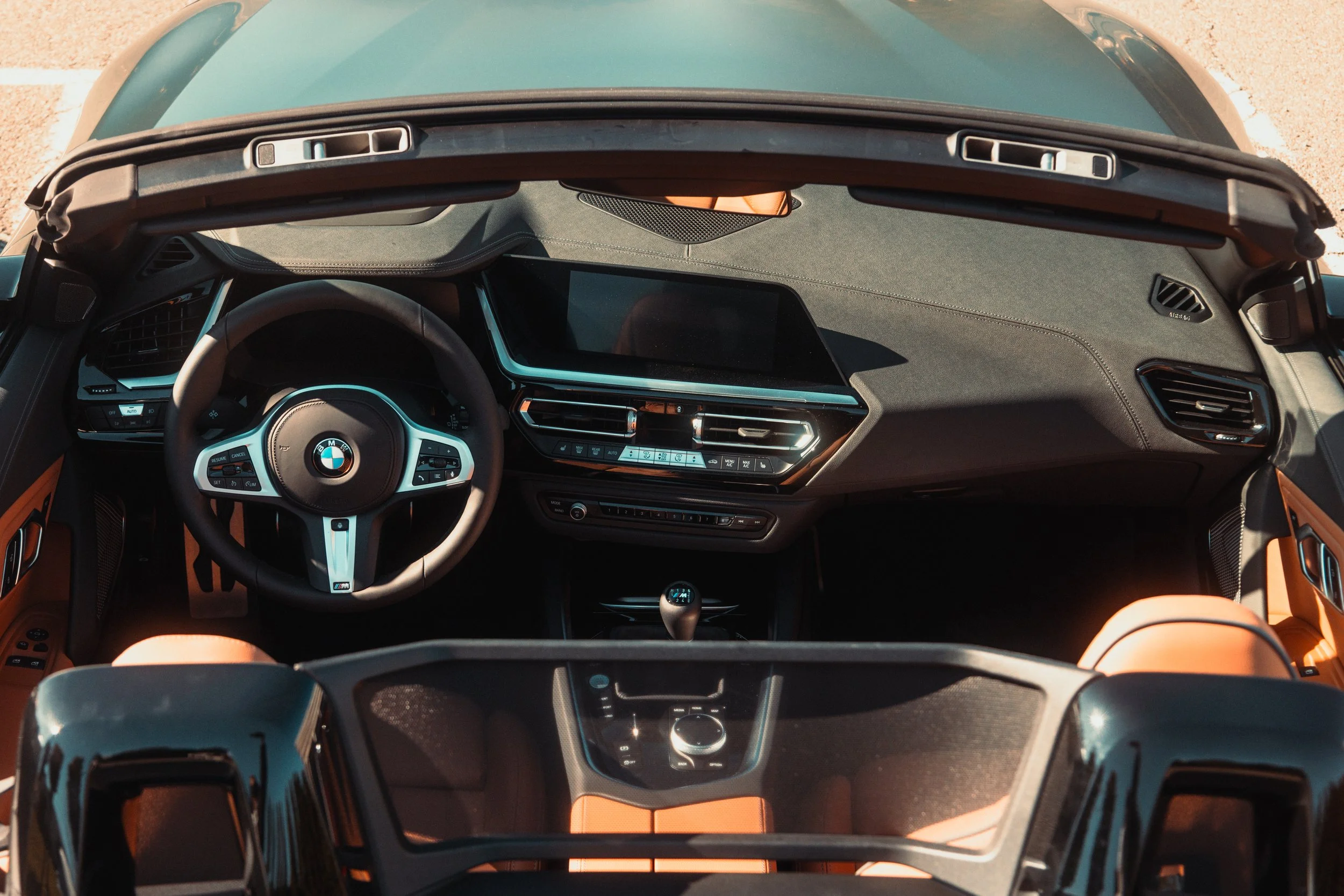 Interior of a BMW convertible with black dashboard, steering wheel, and gear shift, and tan leather seats, taken from above.