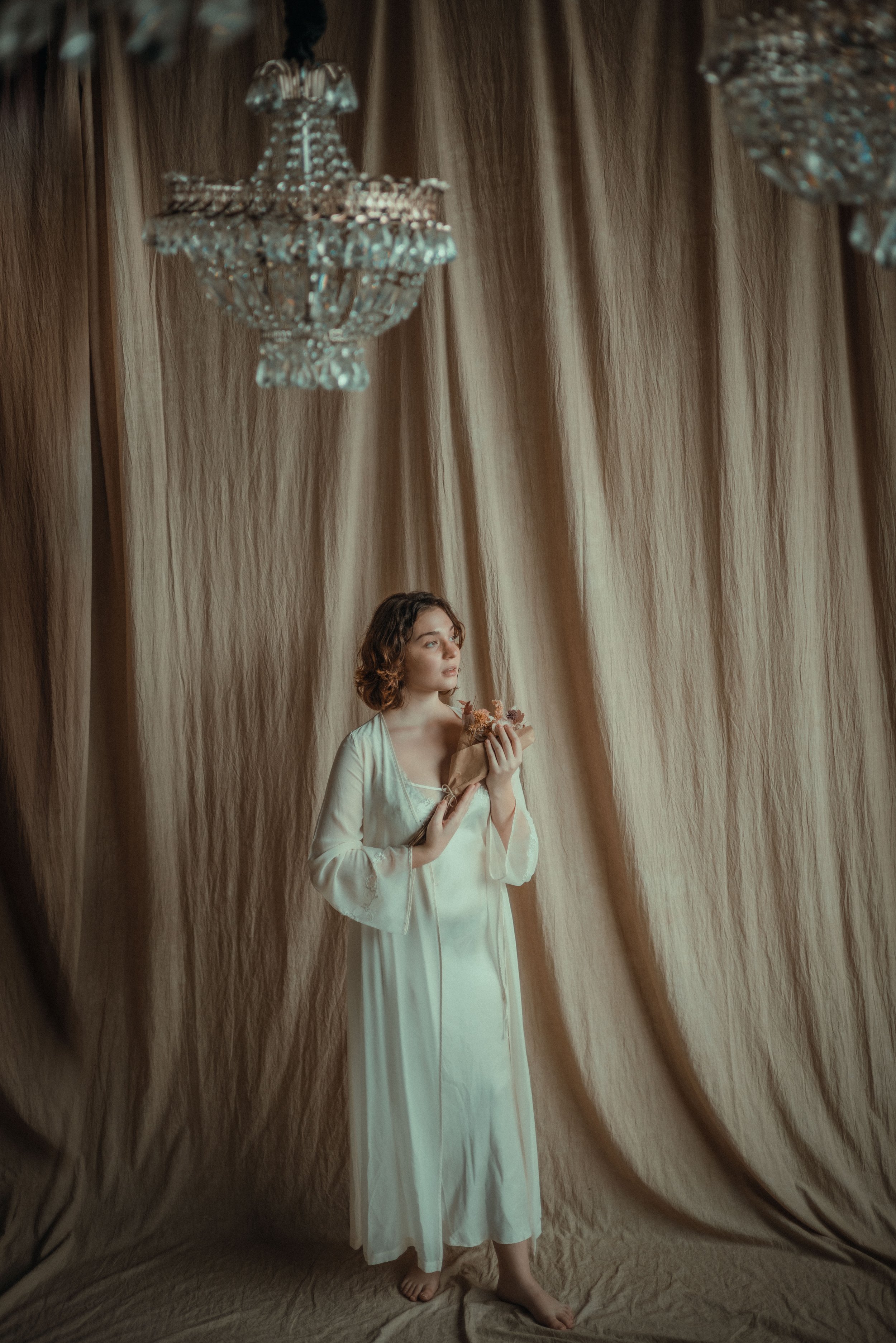 A young woman in a white nightgown stands barefoot before a beige curtain backdrop, holding a bouquet of flowers and gazing thoughtfully upward, with a large crystal chandelier hanging above.