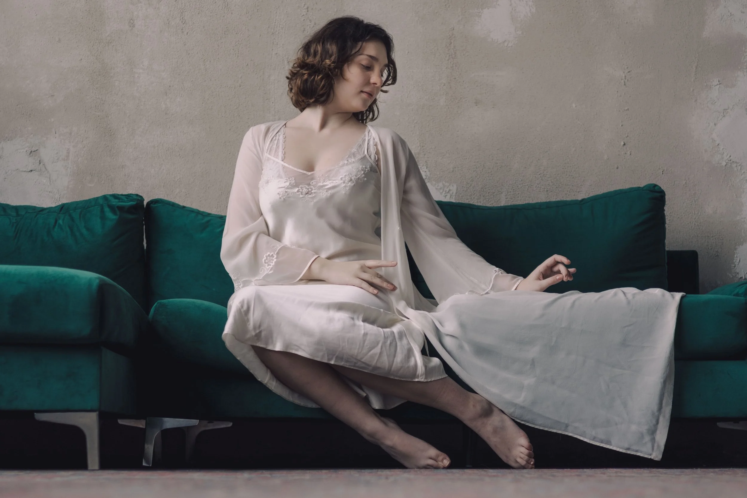 A woman in satin pajamas sitting on a green couch with a distressed wall in the background.