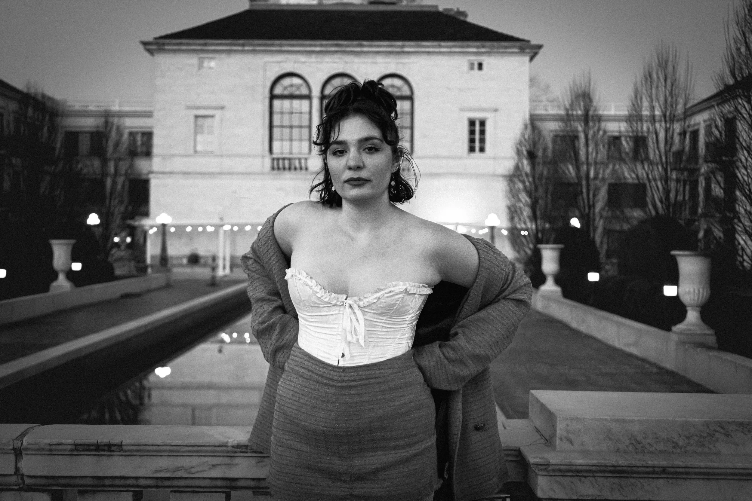 A woman wearing a strapless top with a bow and a sweater is standing outdoors at night in front of a large building, with trees and illuminated lampposts in the background.