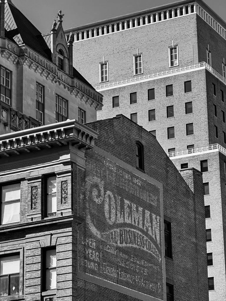 Black and white photo of city buildings, including a historic building with ornate architecture and a large painted advertisement on a brick wall. The advertisement promotes Cold Man Paints and other products, with text partially faded.