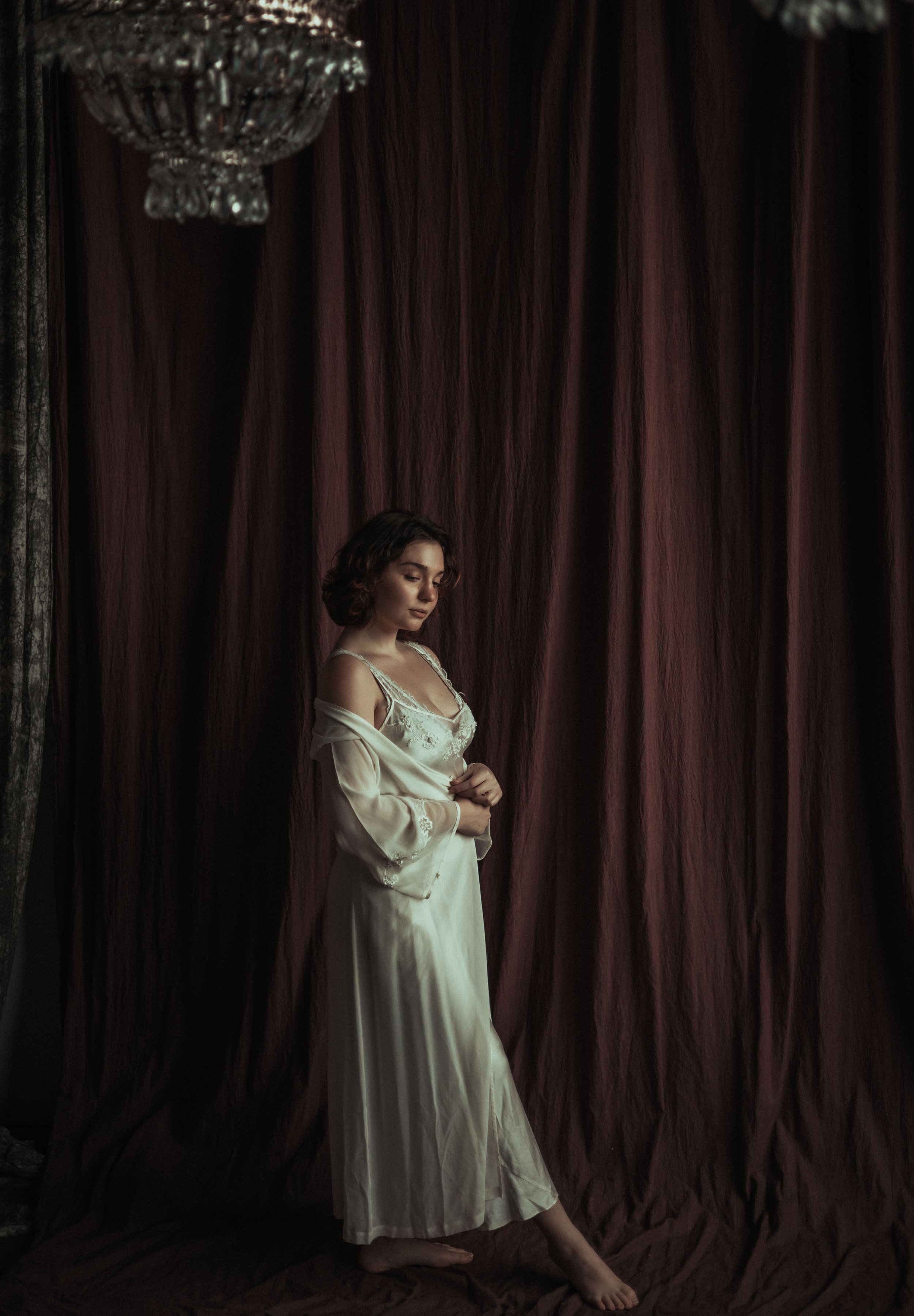 A woman standing barefoot in front of dark red curtains, wearing cream-colored nightwear and a silk robe, with short wavy hair, looking down.