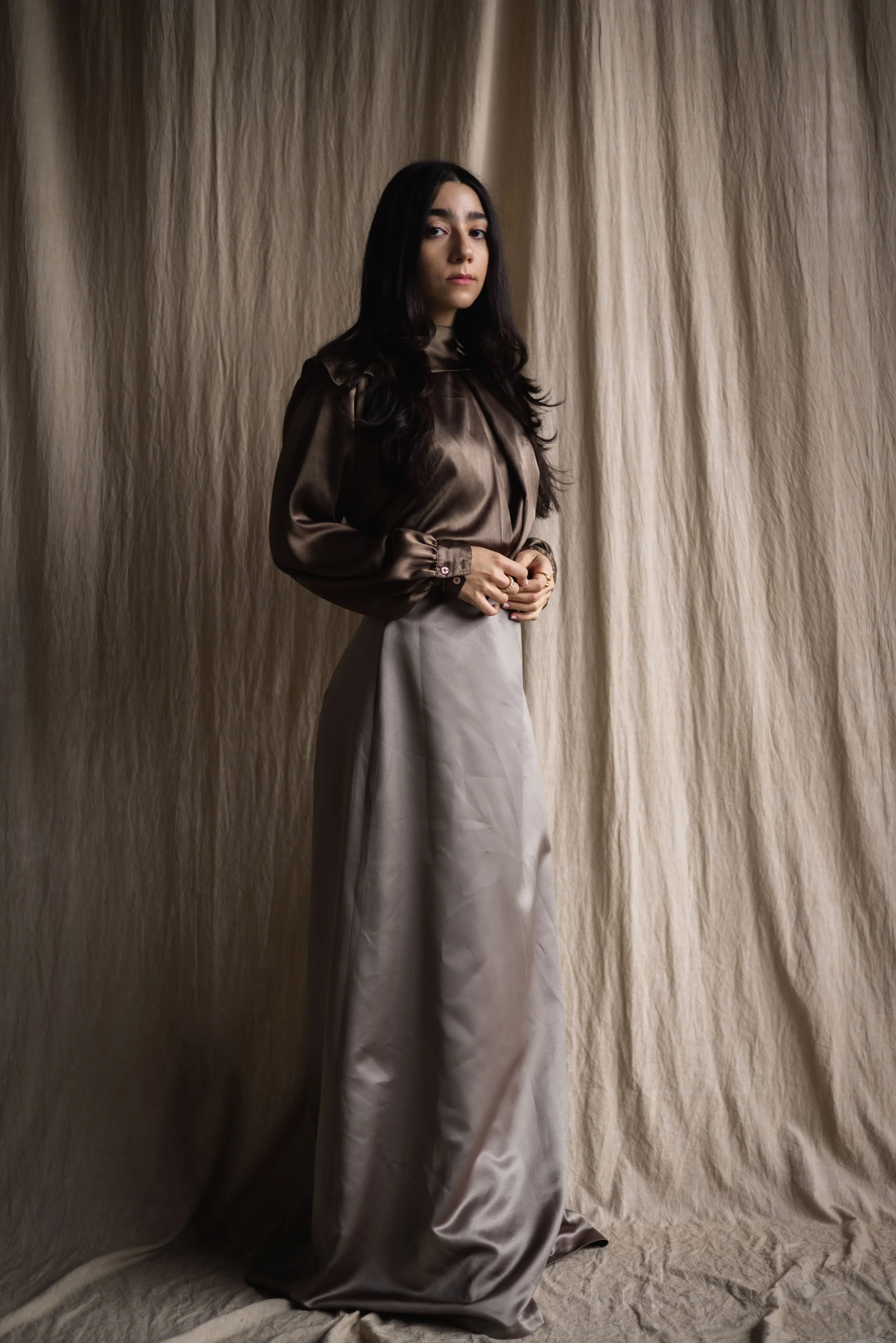 A woman with long dark hair stands in front of beige fabric backdrop, wearing a brown satin blouse and high-waisted beige satin wide-leg pants.
