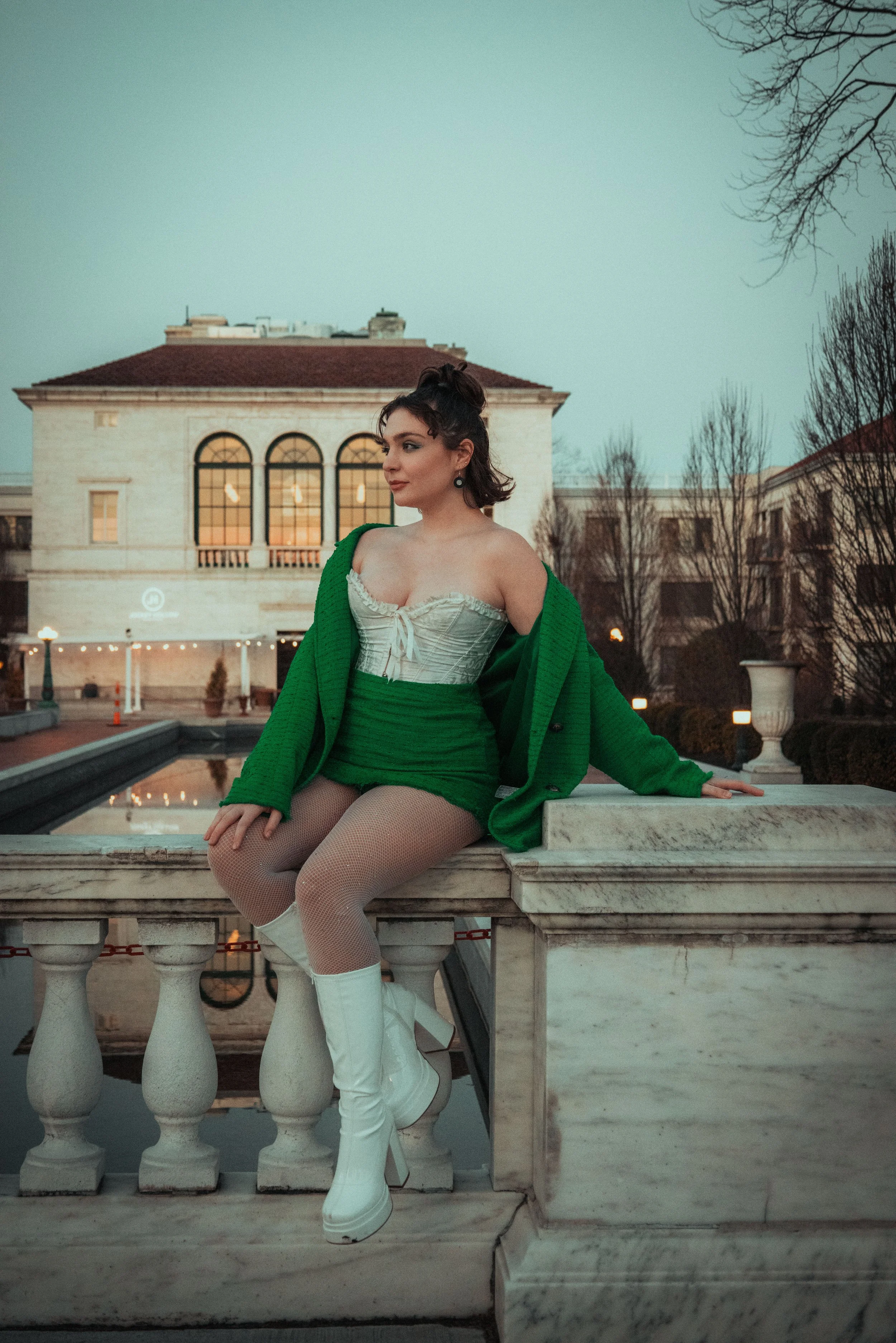 A woman sitting on a stone railing outdoors at dusk, wearing a white corset, green skirt and jacket, fishnet stockings and white knee-high boots, with a building and leafless trees in the background.