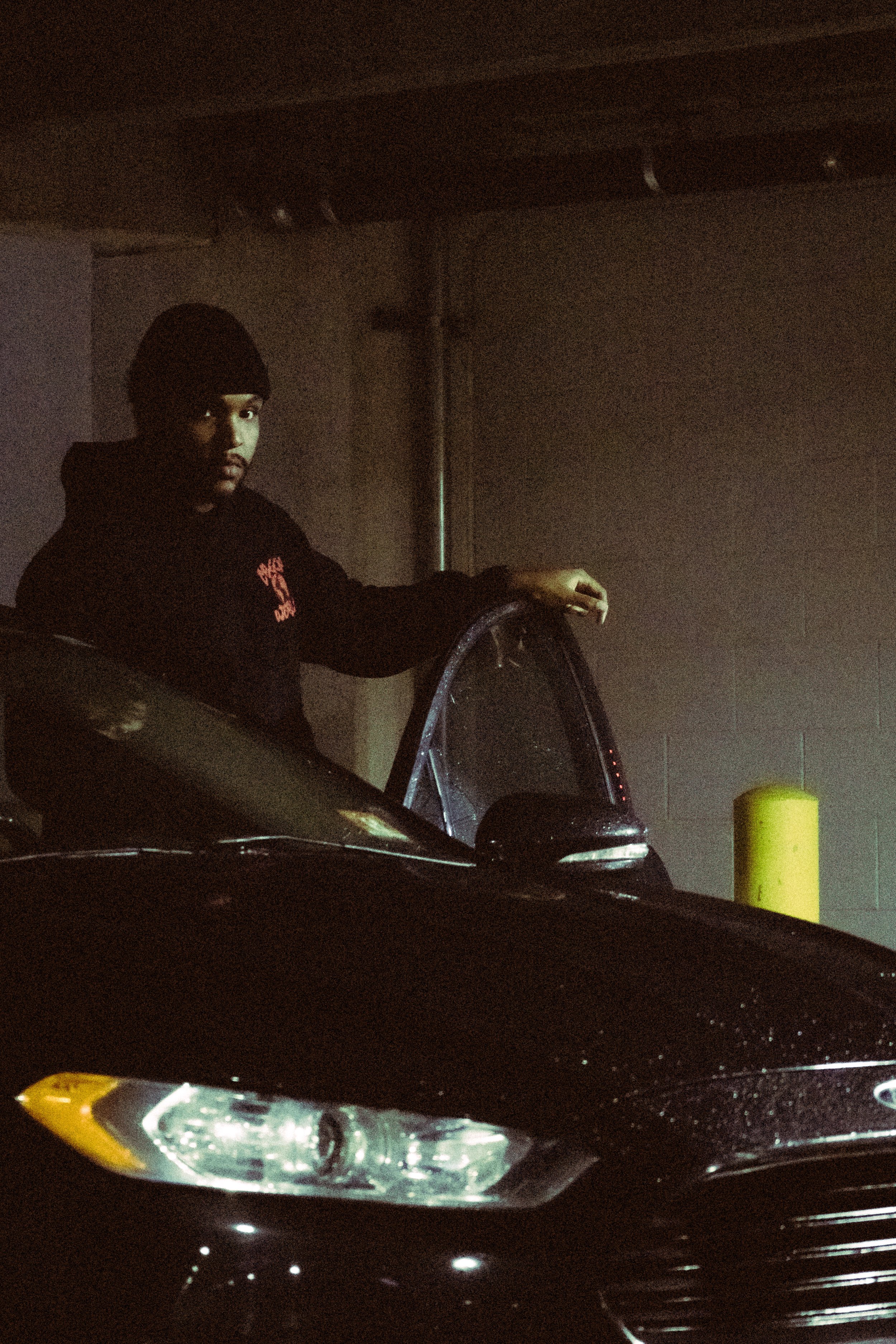 A young man wearing a black hoodie and black beanie standing next to a black car in a dimly lit parking garage.