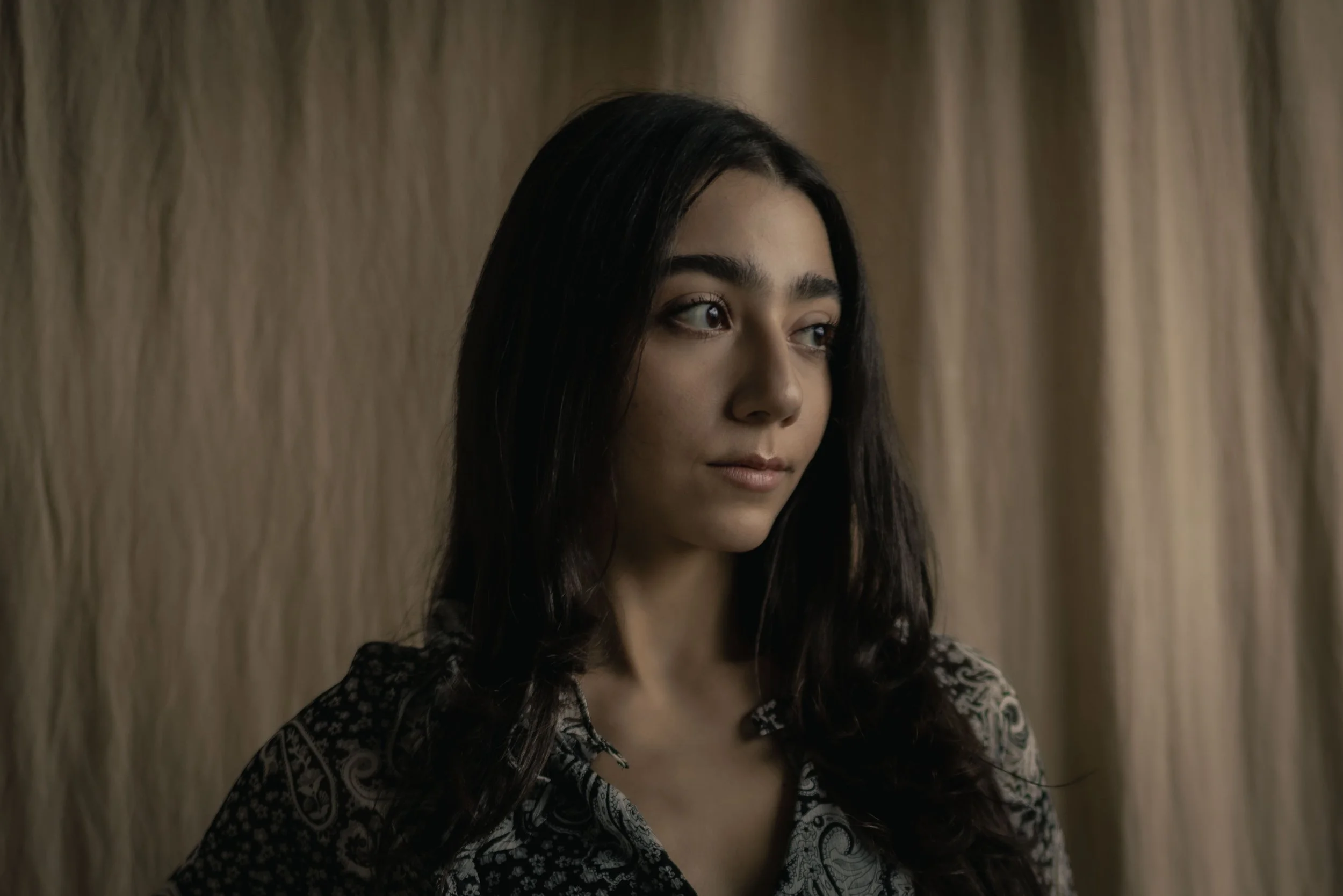 A woman with long dark hair and light skin, looking to the side, standing in front of a beige curtain.