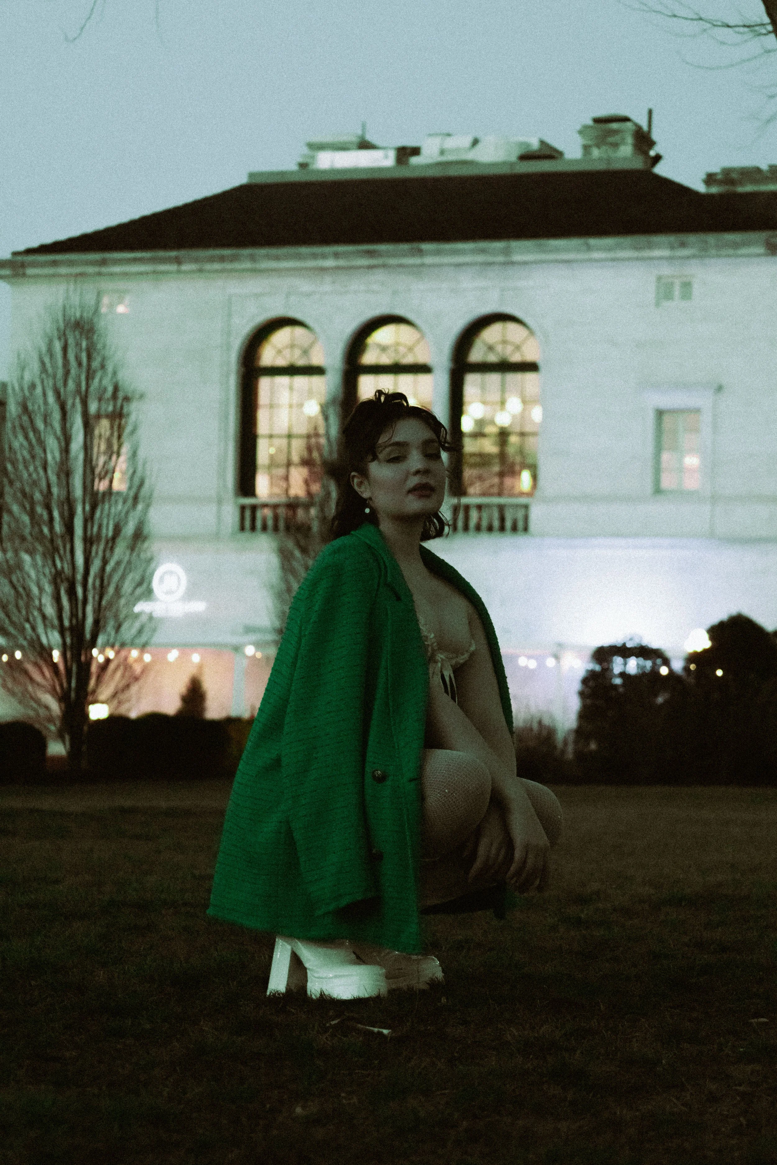 A woman with dark, wavy hair and makeup, wearing a green coat, is outdoors at night with city lights and a white building with arched windows behind her.