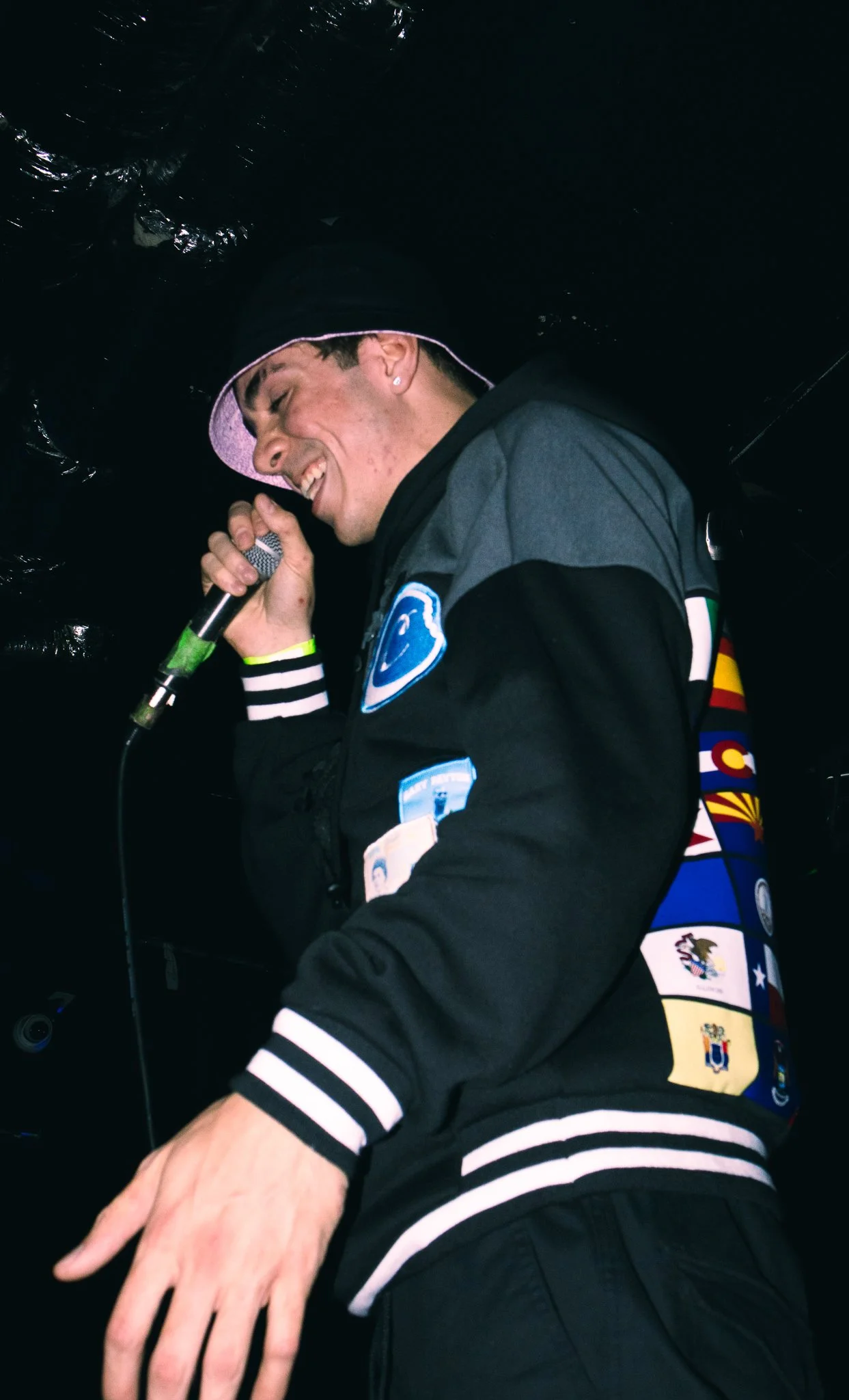 Young man singing into a microphone, wearing a black jacket with patches and a pink cap, smiling with eyes closed, against a dark background.