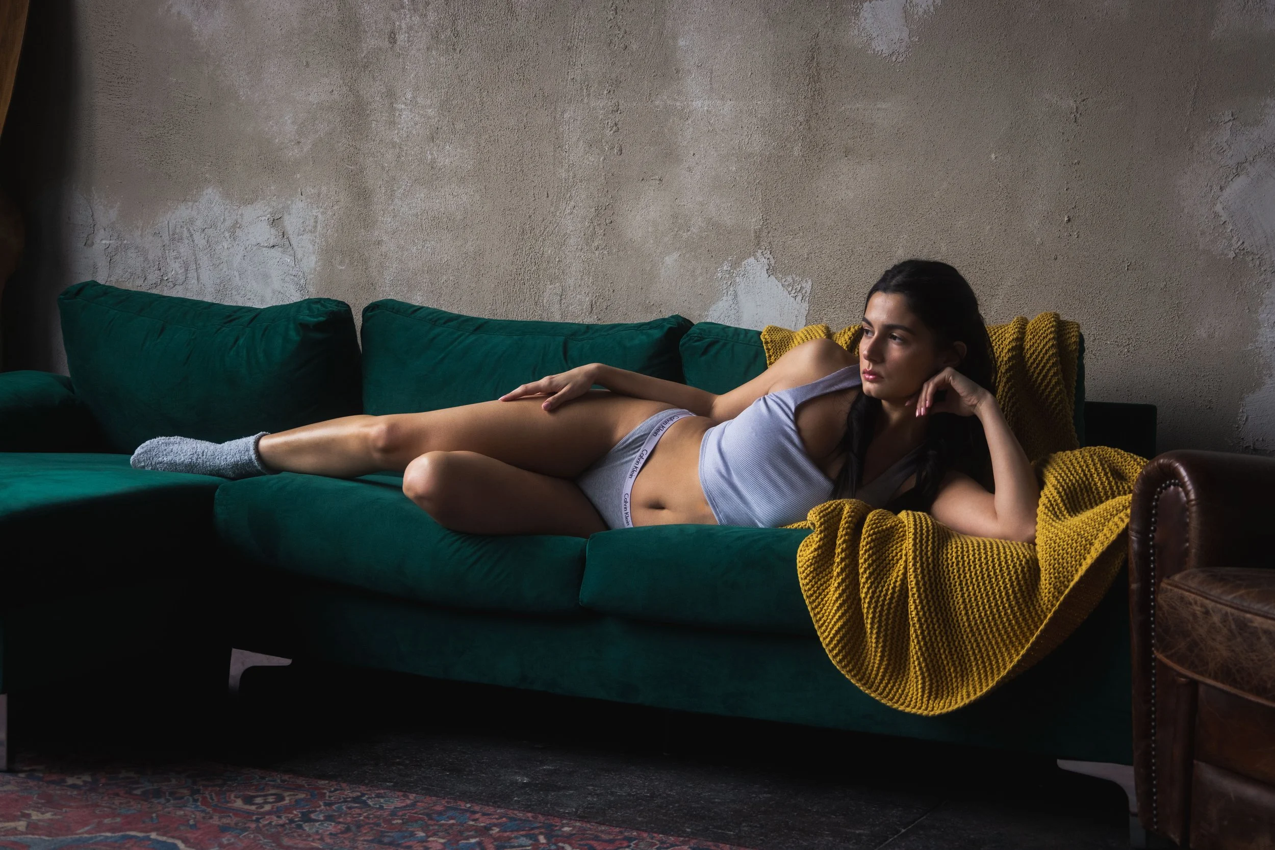 A woman lying on a green sofa in a relaxed pose, wearing a gray tank top, gray underwear, and gray socks, with a yellow knitted blanket draped over the armrest in a room with a textured wall.