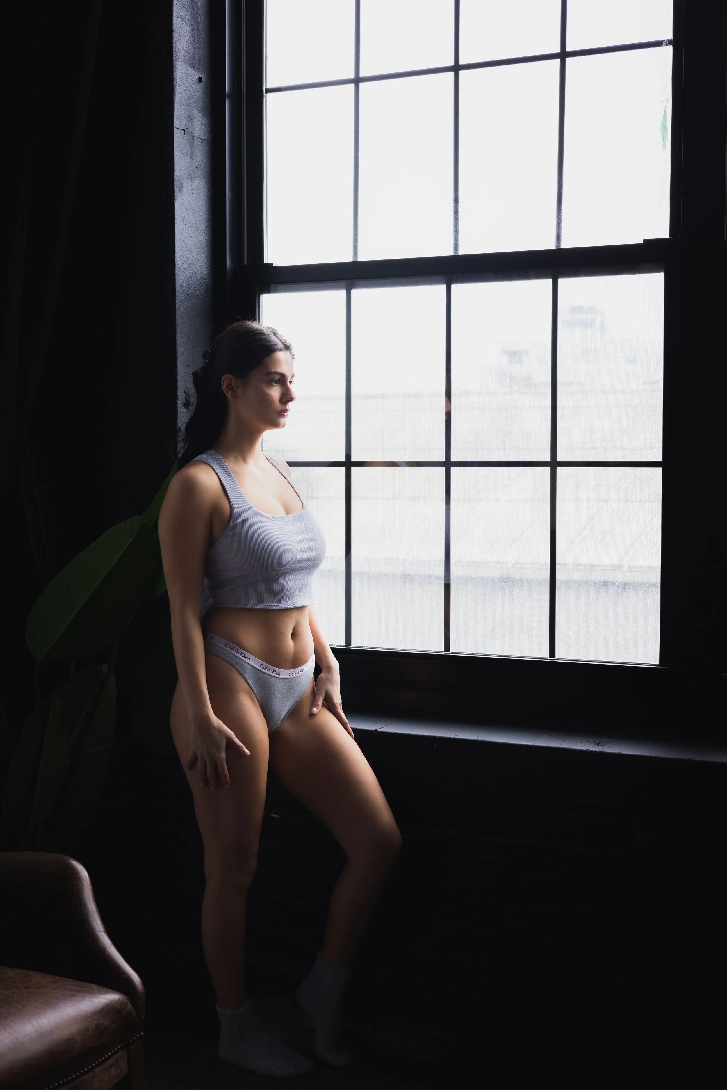 A woman in gray underwear and a gray tank top standing by a large window, looking outside, with natural light illuminating her profile, in an industrial-style room.