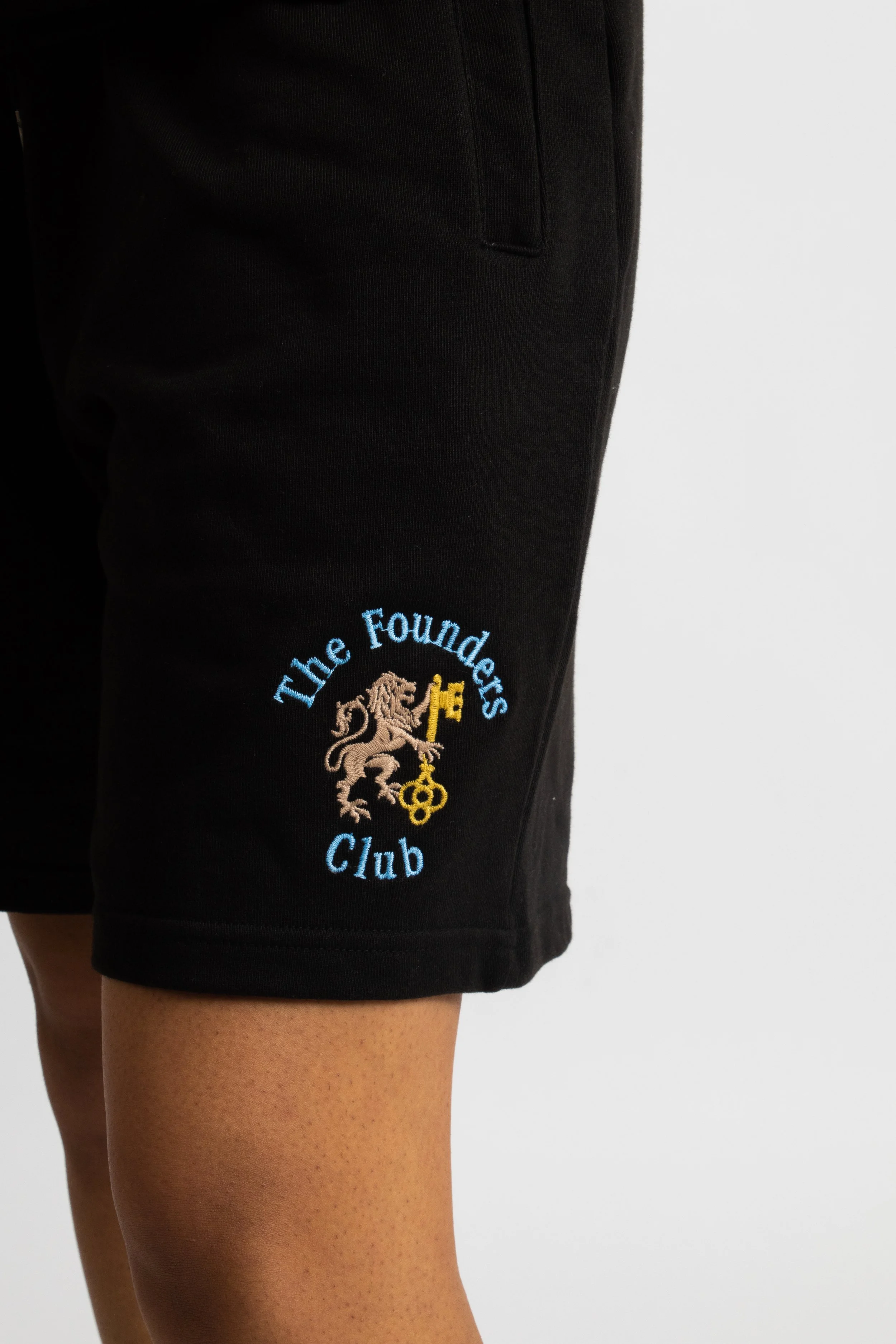 Close-up of a person wearing black shorts with an embroidered logo of a lion holding a key and the text 'The Founders Club' on the side.