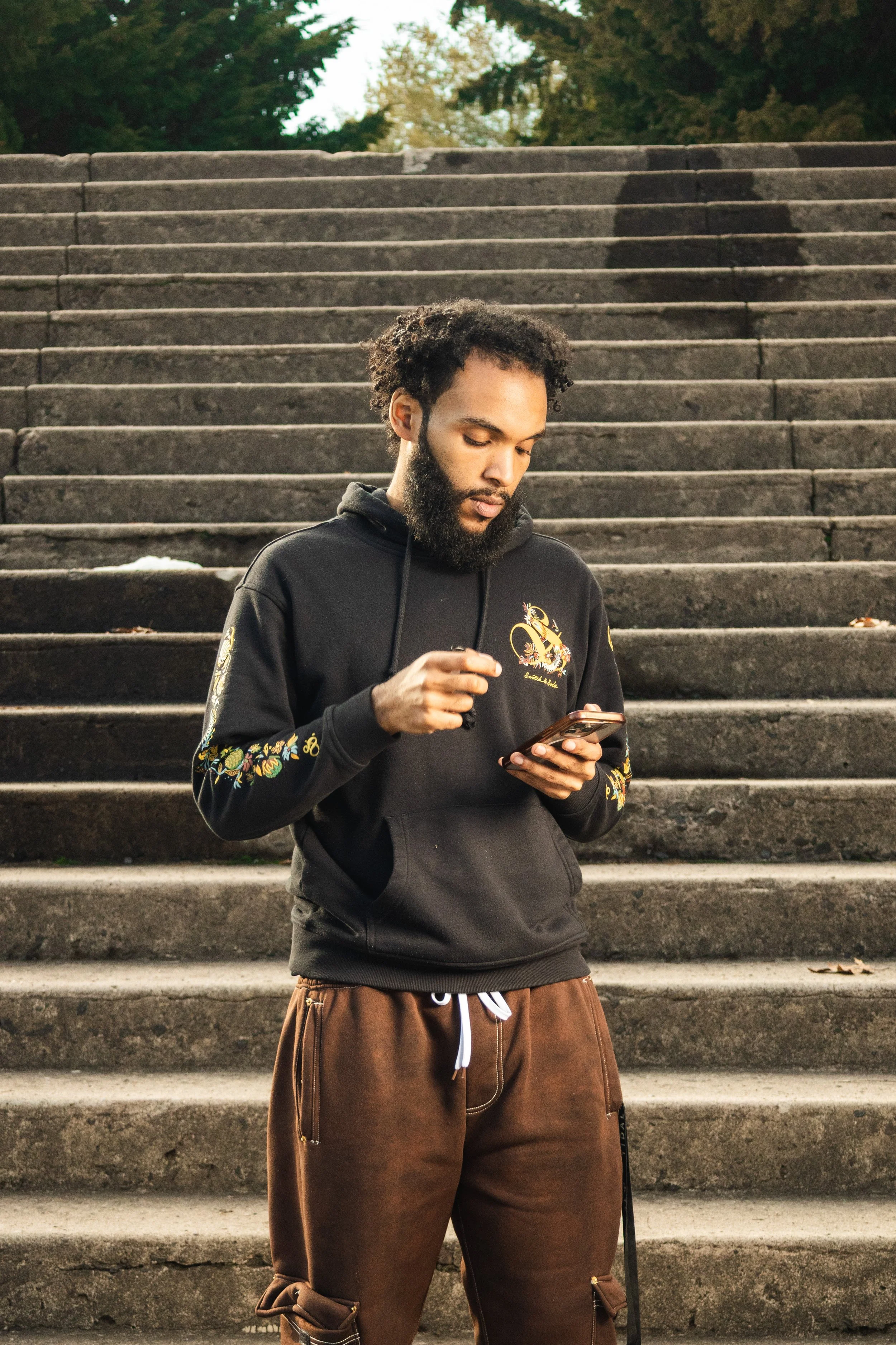 A young man with a beard and curly hair is standing on outdoor stairs, looking at his phone while holding earbuds.