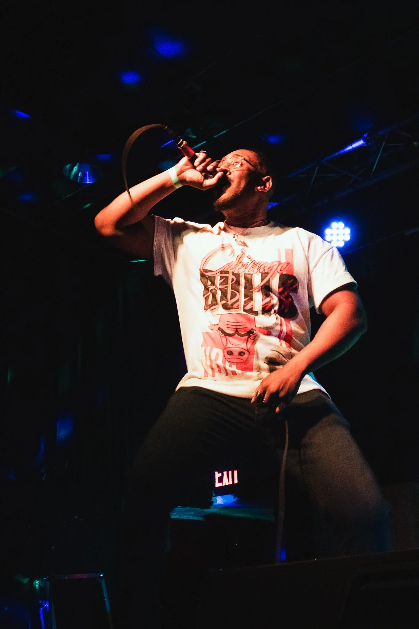 A man singing into a microphone on stage in a dimly lit setting with blue lights, wearing a Chicago Bulls t-shirt and black pants.