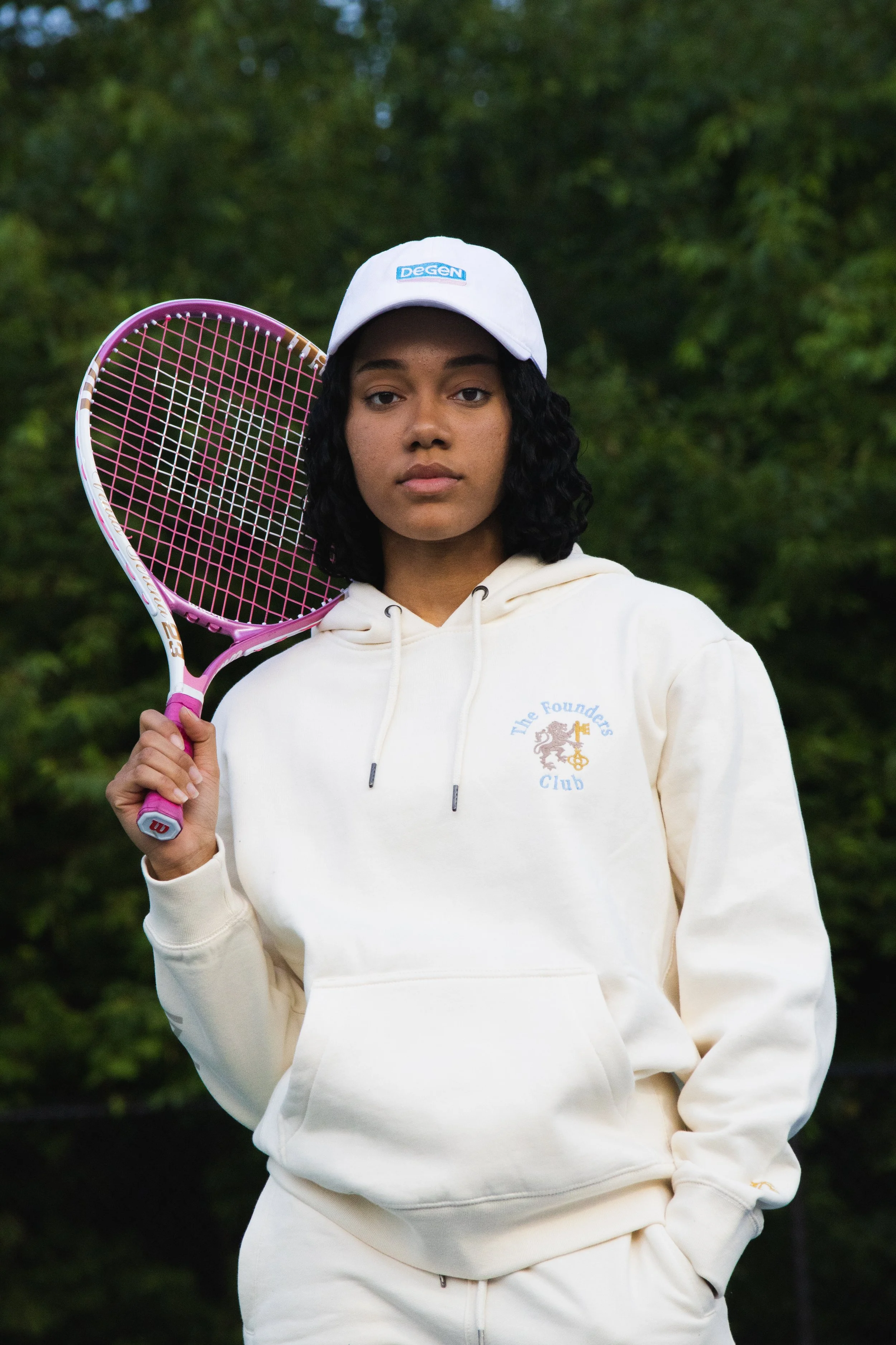 A young woman with dark, curly hair wearing a white hoodie and white cap, holding a pink tennis racket on her right shoulder, standing outdoors against a backdrop of green trees.