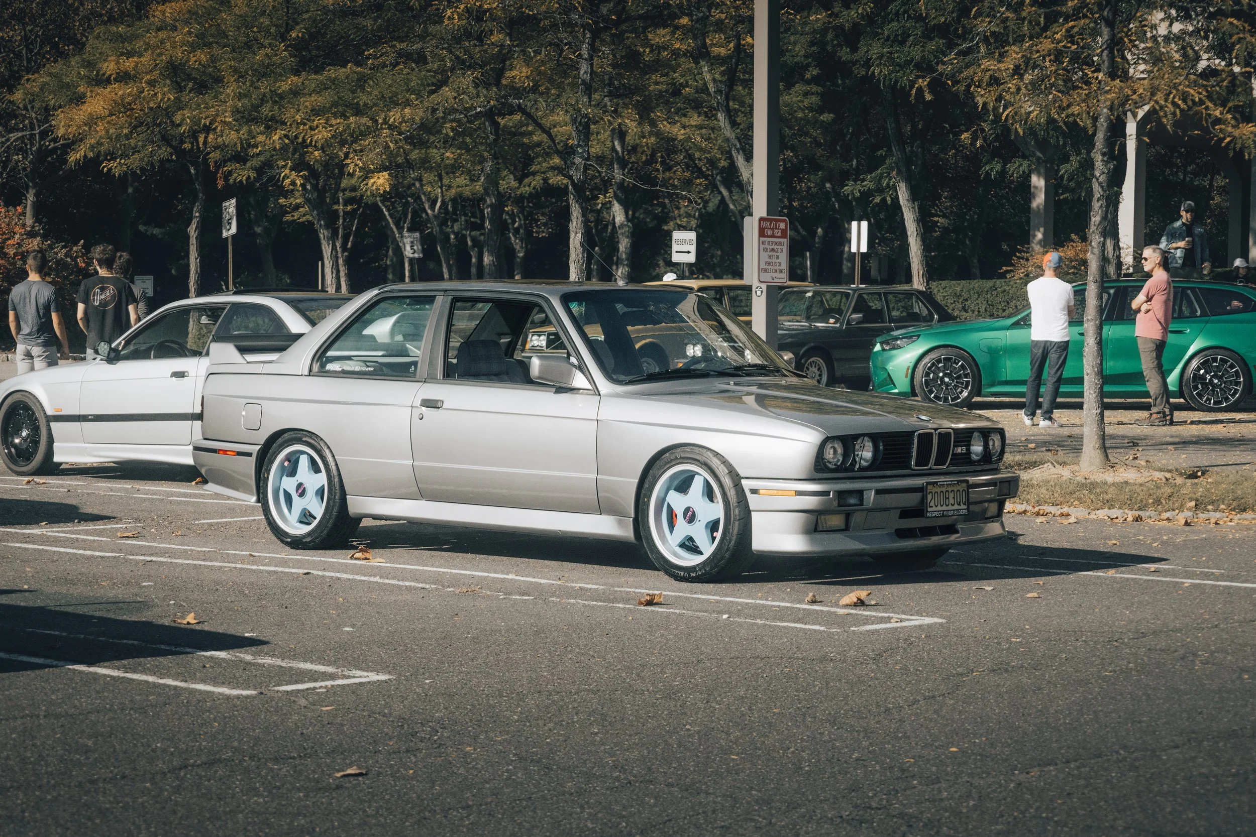 A silver vintage car parked in a parking lot, with other cars and people standing around in the background, trees and autumn leaves on the ground.