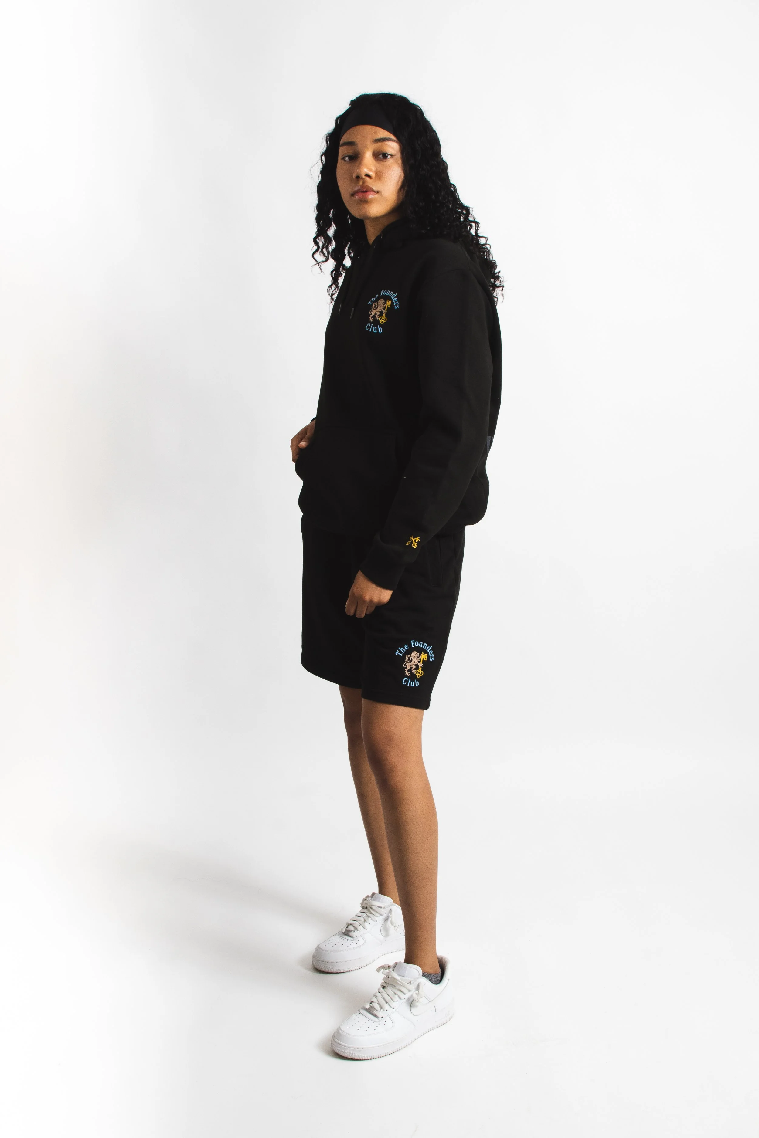 Young woman with curly hair wearing a black hoodie and shorts, branded with 'The Founders Club' logo, and white sneakers, standing against a white background.