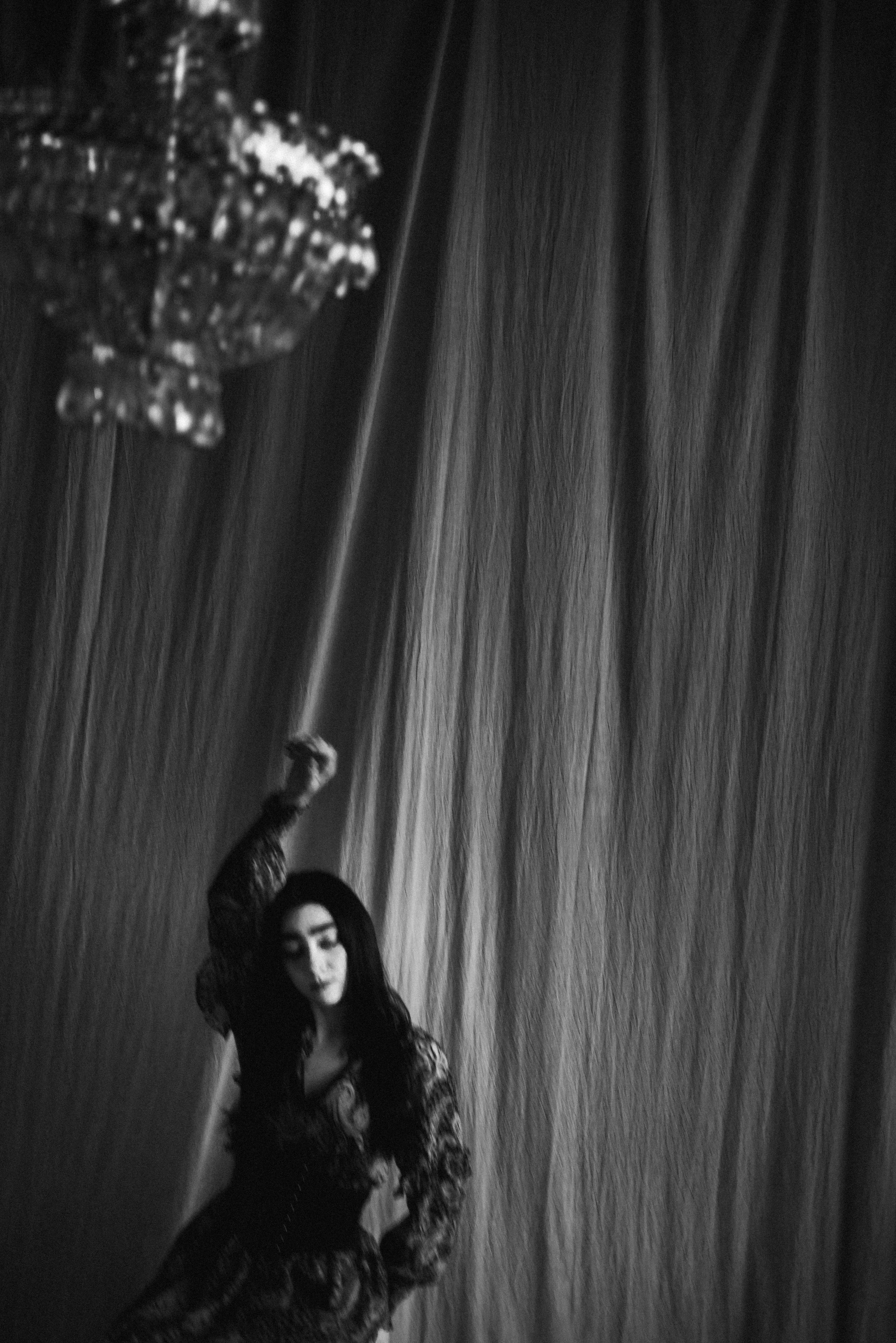 A woman with long dark hair, dressed in dark clothing, standing with one hand in her pocket and the other arm raised. She is in front of a textured curtain, with a blurred decorative object in the background.