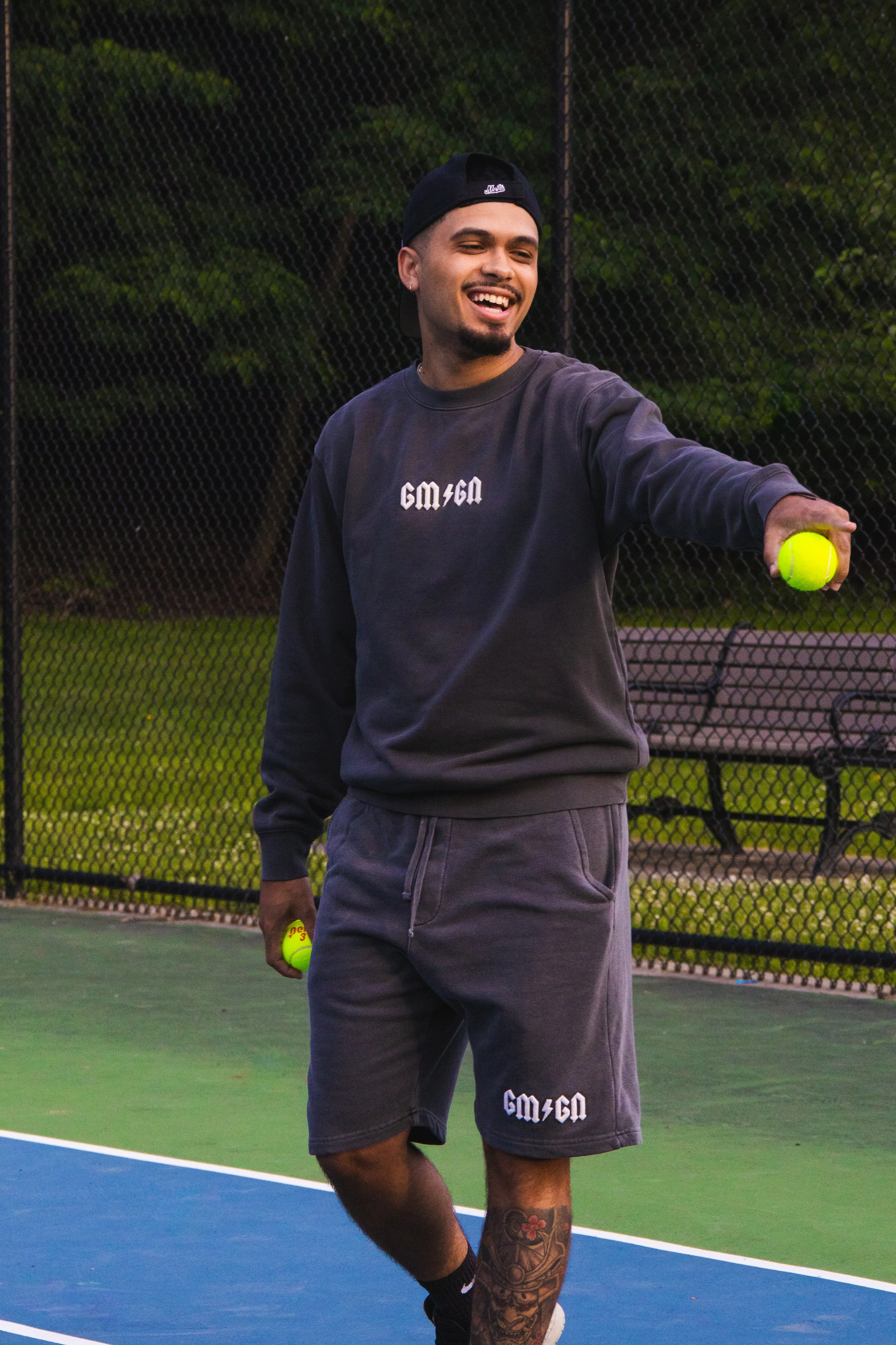 A young man smiling and playing tennis on a court, holding a tennis ball and wearing casual sportswear and a baseball cap.