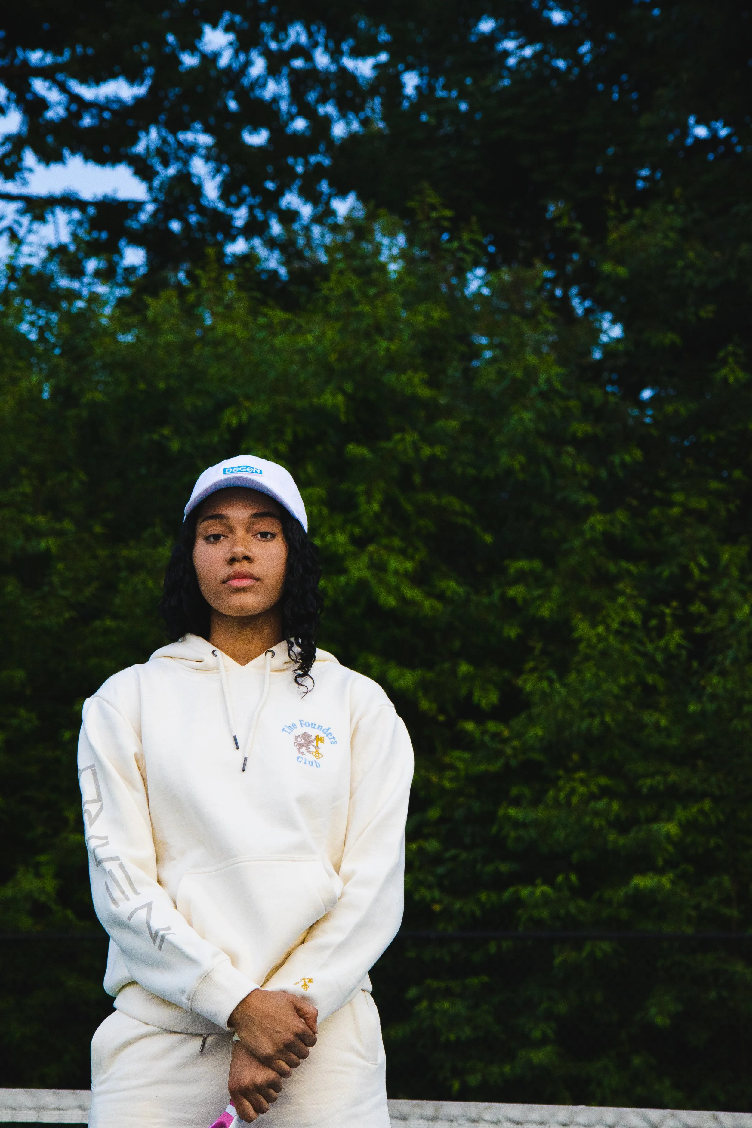 A young woman with curly black hair stands outdoors in front of a forested background. She wears a white hoodie with a small logo on the chest, a white baseball cap, and beige pants, with her hands clasped in front of her.