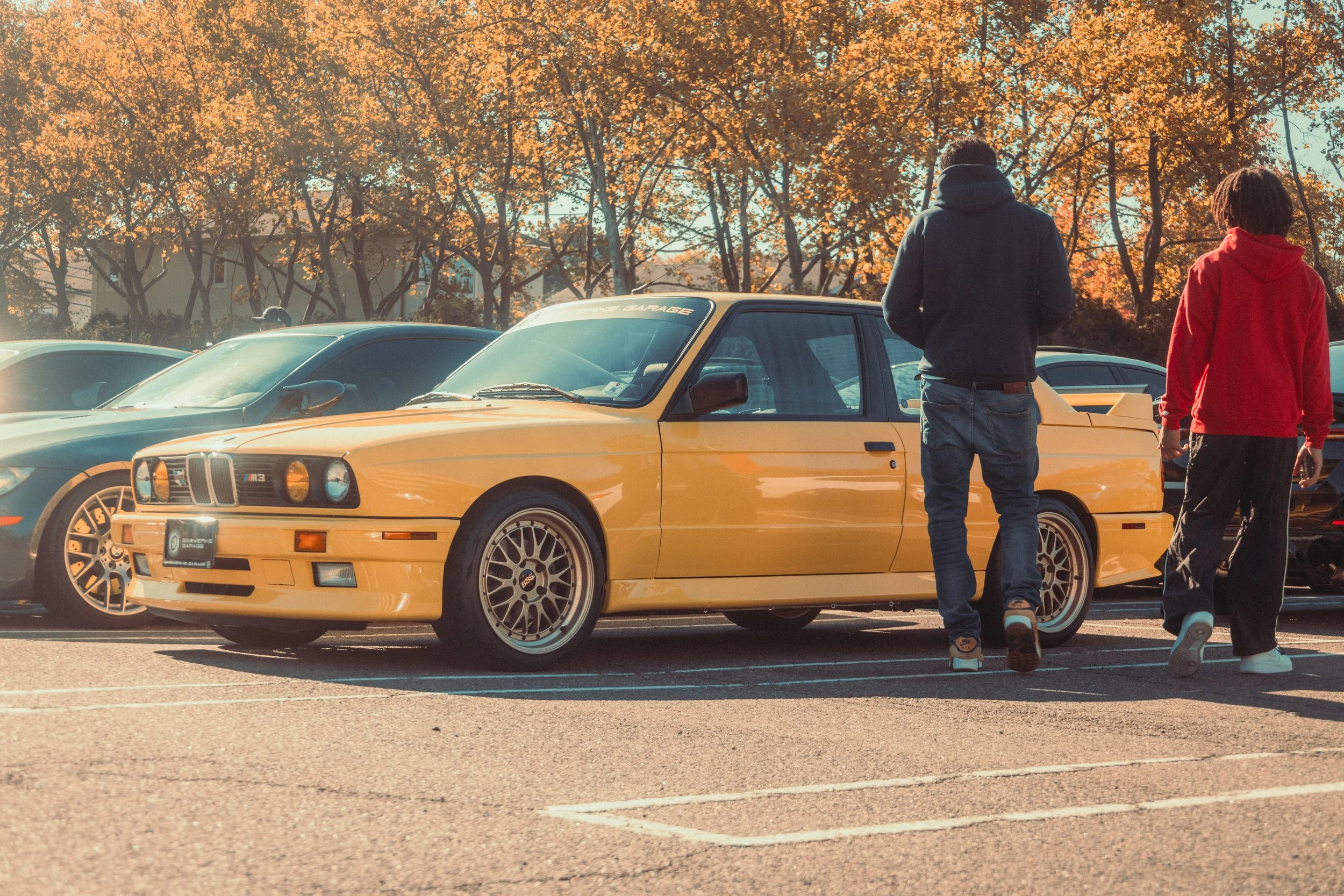 A yellow vintage BMW M3 parked in a lot during fall, with two people walking nearby and trees with orange leaves in the background.