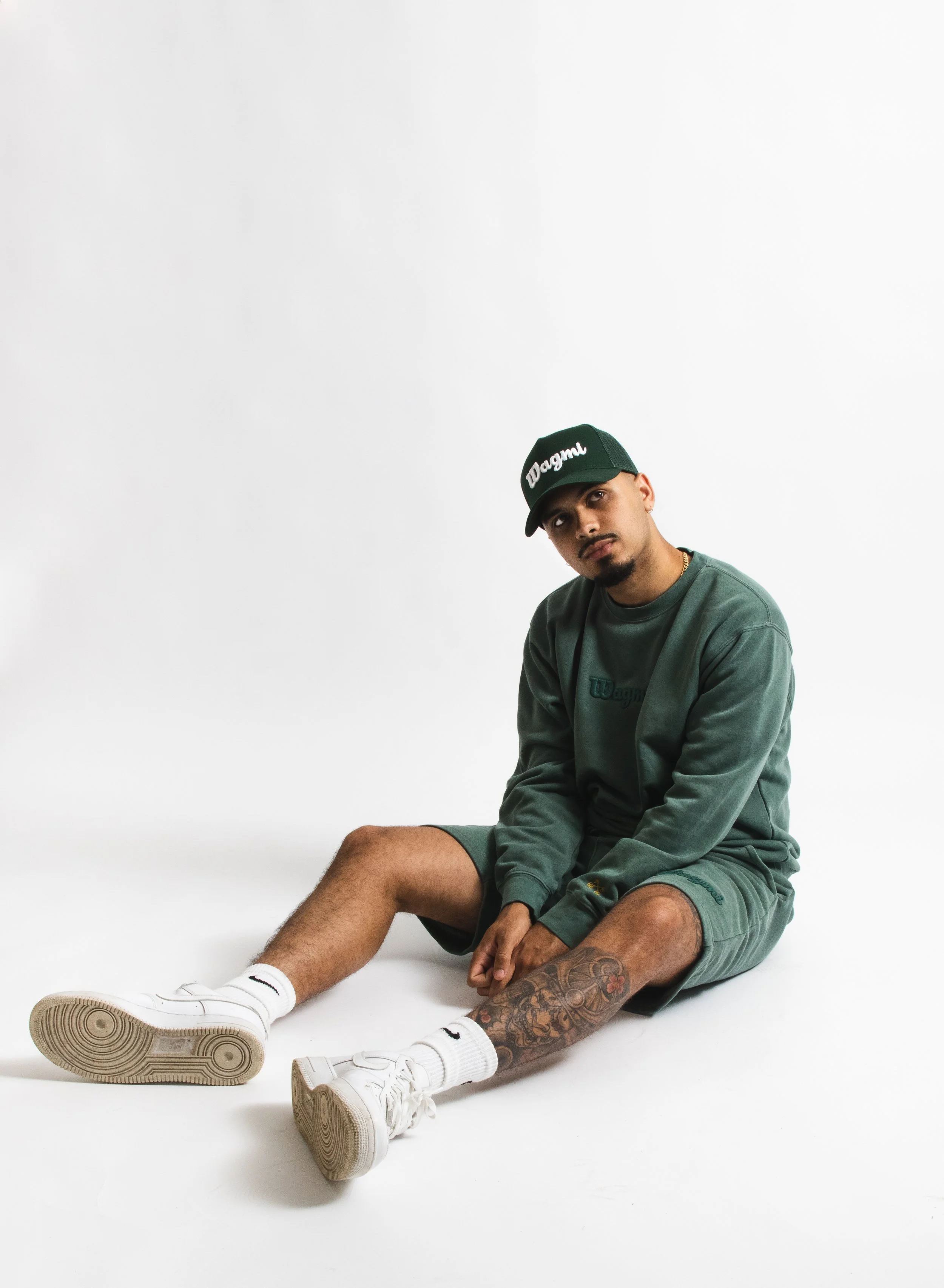 A young man sitting on the floor against a plain white background, wearing a green sweatshirt, green shorts, white sneakers, white socks, and a green baseball cap with 'Dagma' embroidered on it, looking at the camera.