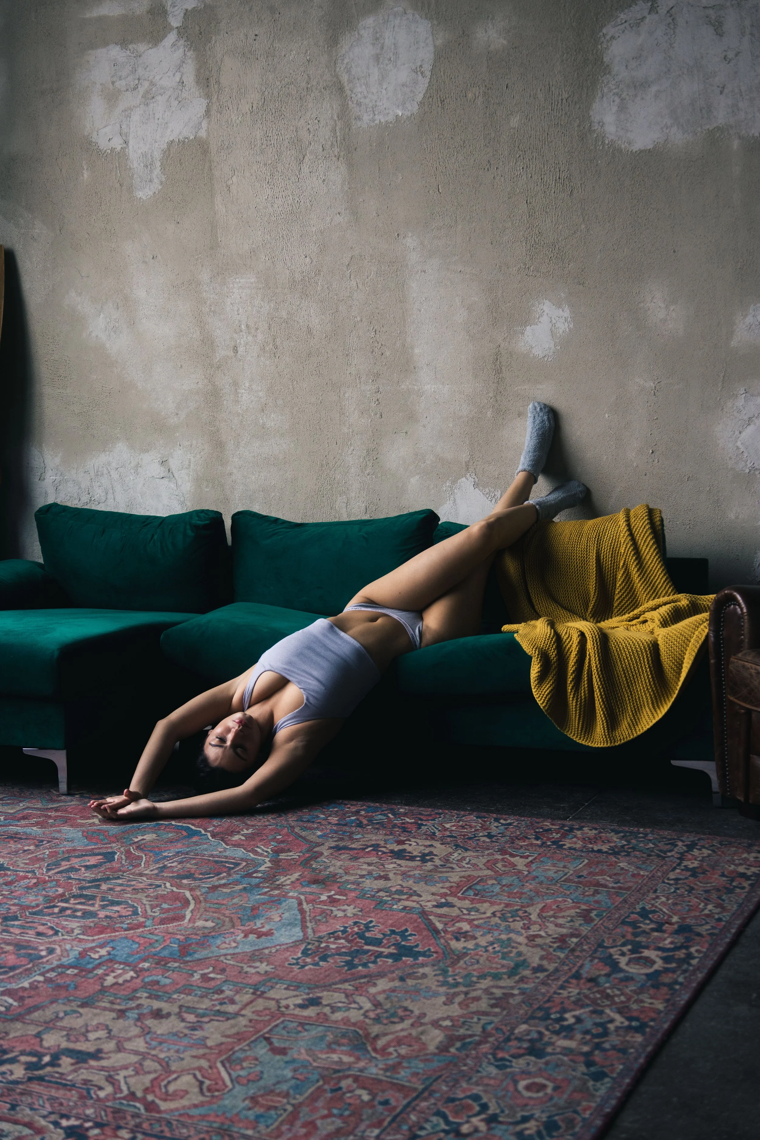 Woman lying on a green sofa with her legs propped against the wall, wearing gray underwear and a tank top, in a room with a distressed wall and a patterned rug.