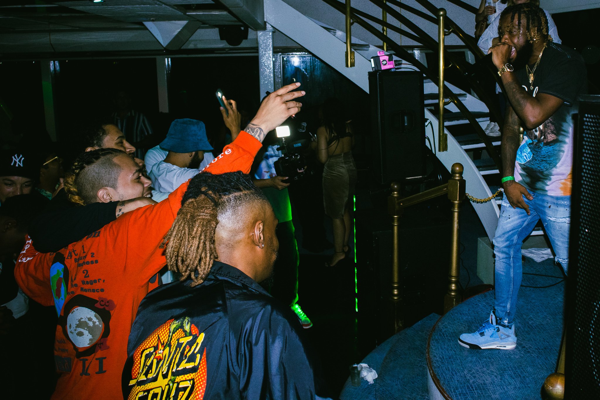 A man performing on stage in a nightclub, wearing a black shirt and blue tie-dye jeans, with gold chains and bracelets, in front of a small crowd of fans.