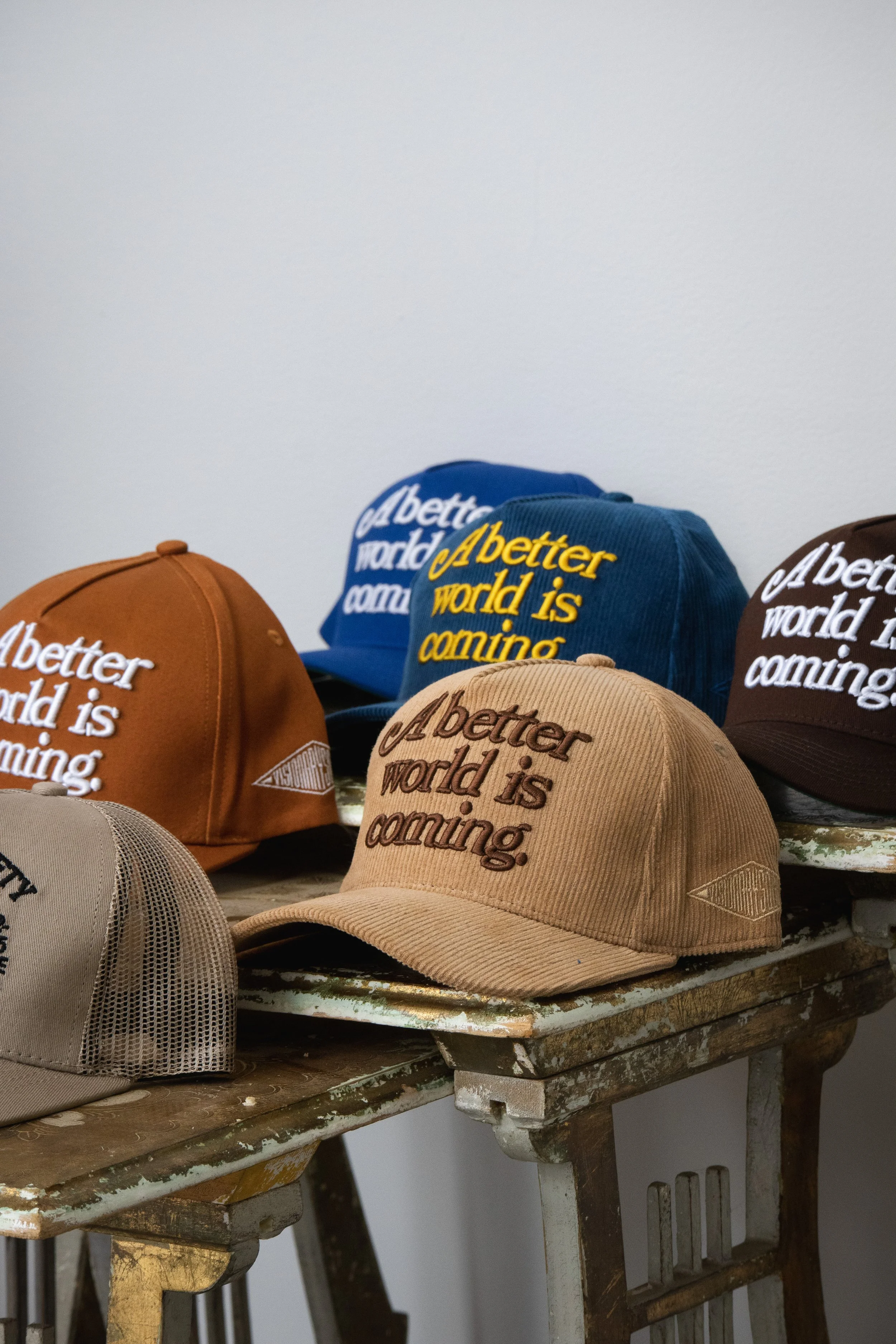 Several baseball caps in various colors with the embroidered message 'A better world is coming' displayed on a rustic wooden table against a plain white wall.