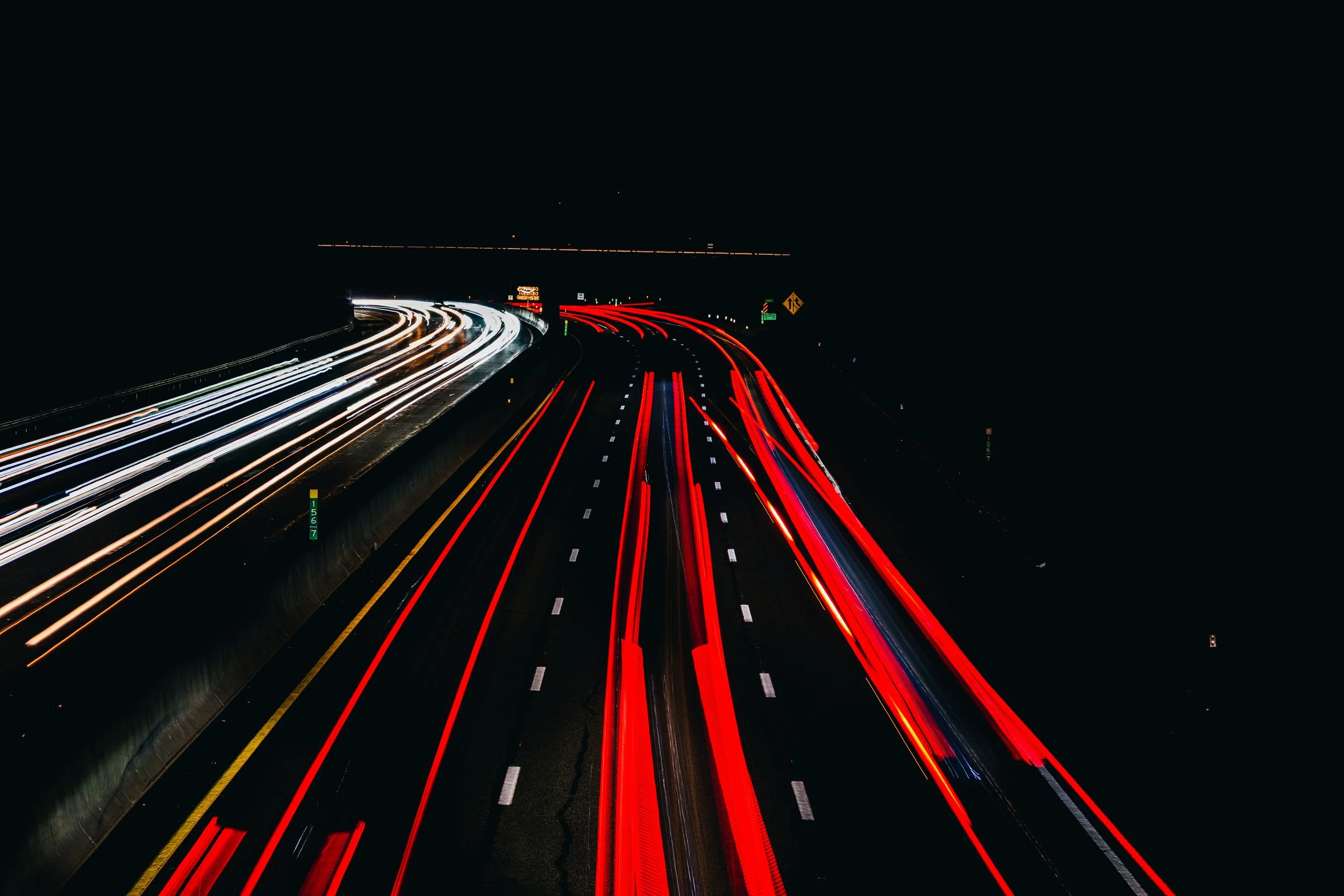 Long exposure photo of a busy highway at night with streaks of white and red lights from moving vehicles.