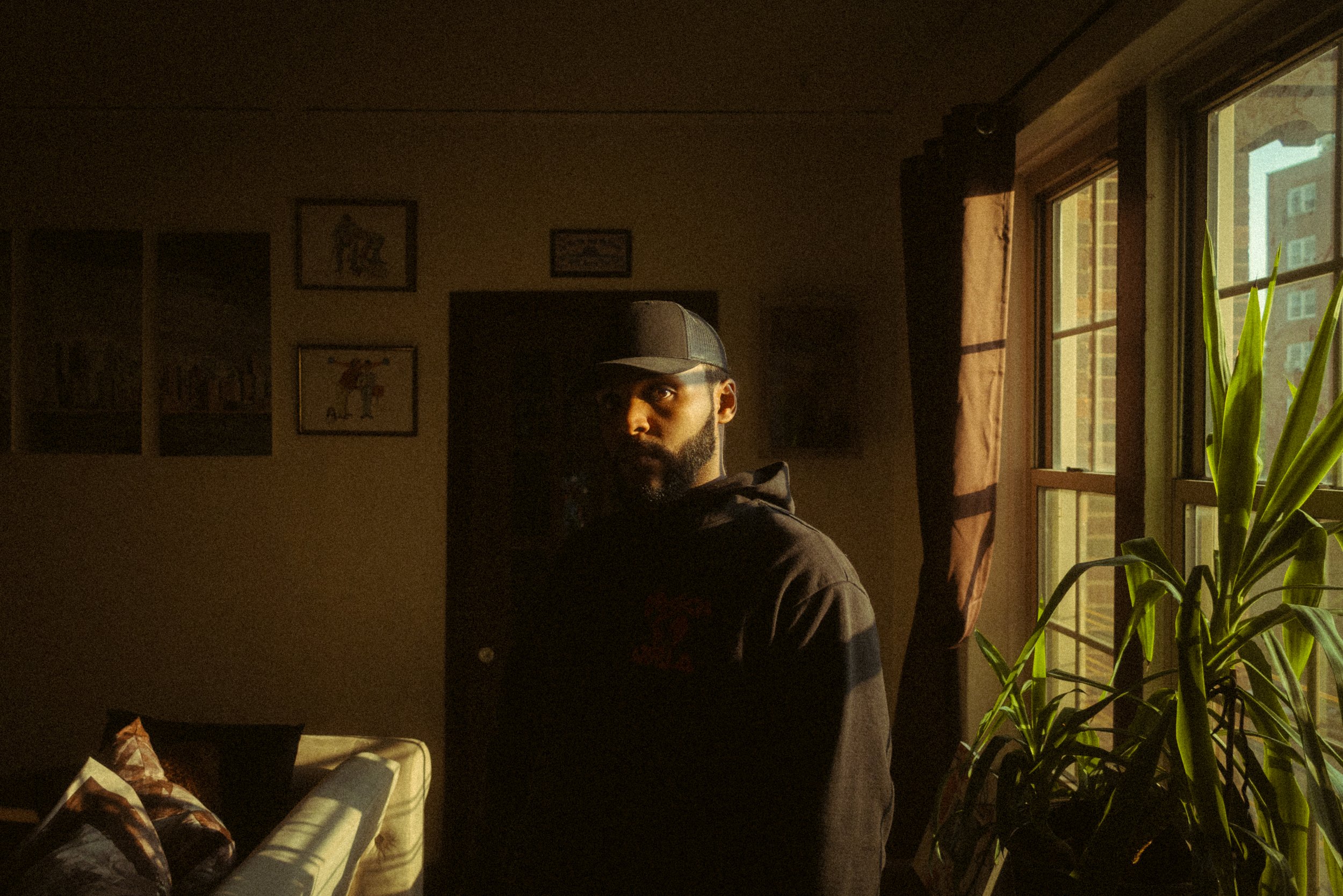 Man with a beard and hat standing near window with sunlight, plants, and framed pictures in background.