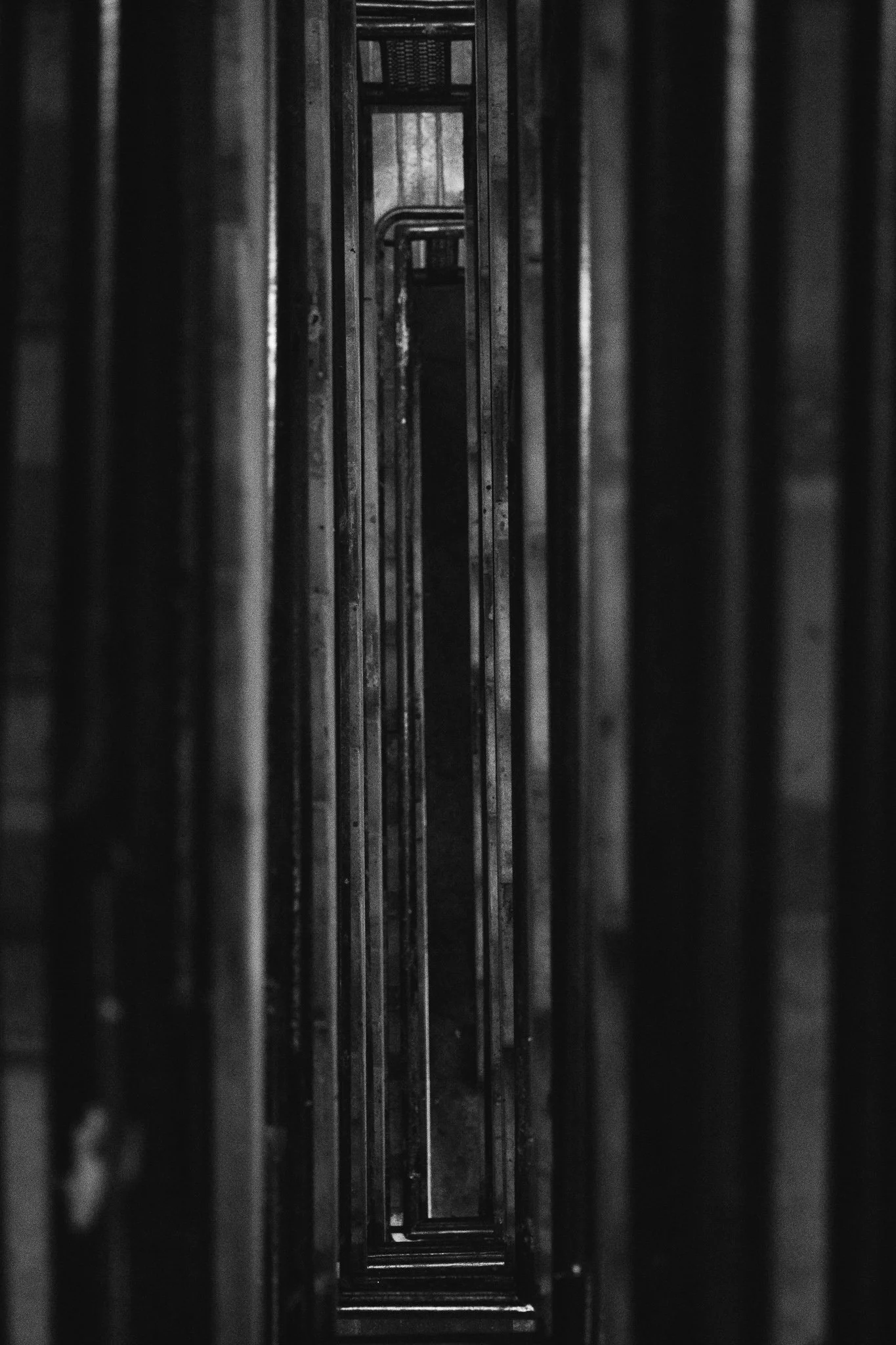 Black and white photo of tall, narrow metallic structures or shelving units seen from above, creating a tunnel-like perspective.