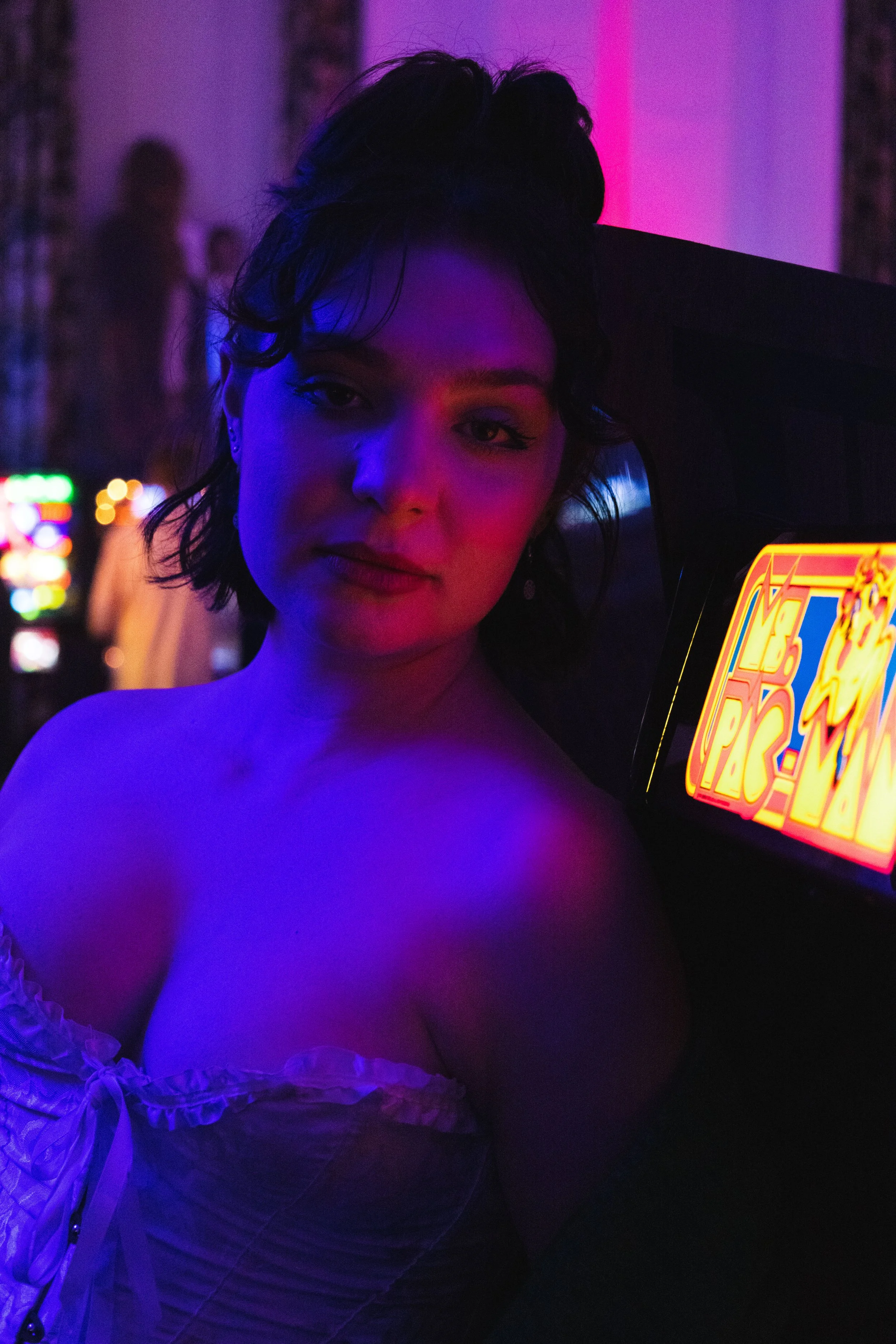 A woman with short dark hair and earrings, illuminated by neon pink and purple lighting, standing next to a brightly lit game machine in a dimly lit room with blurred figures in the background.