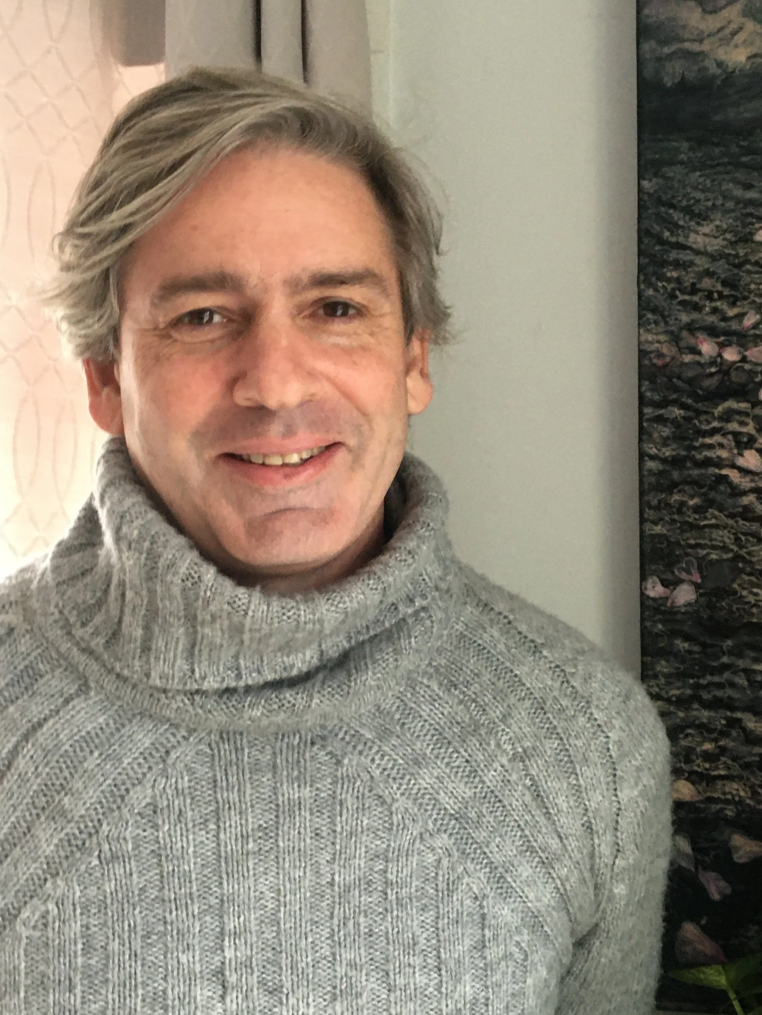 A middle-aged man with gray hair smiling, wearing a gray turtleneck sweater, standing indoors next to a wall with artwork.